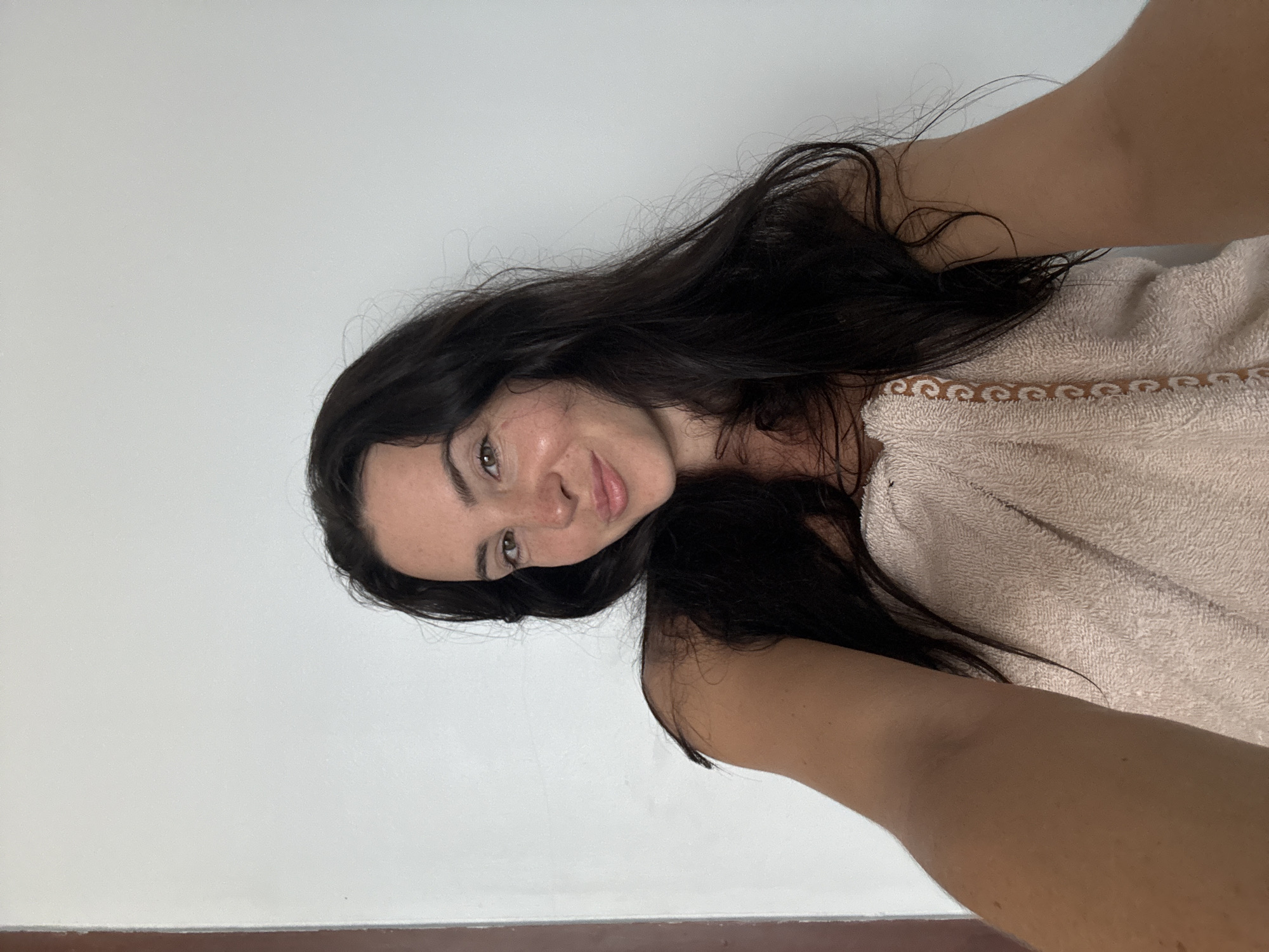 A young woman with long, dark hair is taking a selfie in a room with a white wall and a brown door frame. She is wearing a light pink towel wrapped around her body. She has a slight smile and her eyes are looking towards the camera. Her arm is extended to take the photo. The setting appears to be residential, possibly a hotel room in North Kuta, Indonesia.