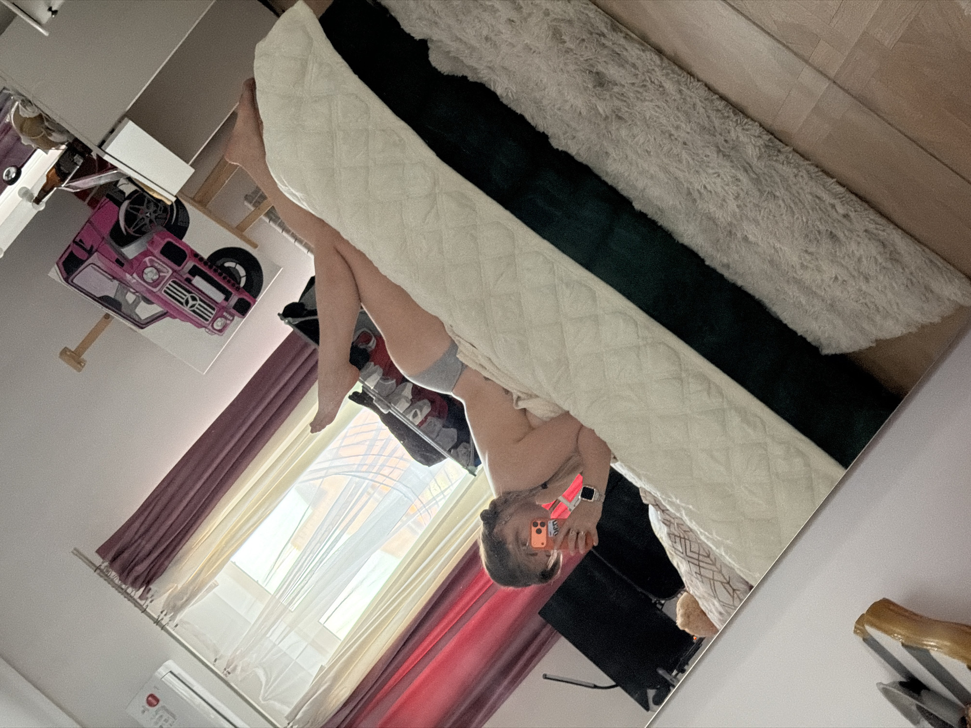 The image appears to be a mirror selfie taken in an indoor environment, likely a bedroom. The reflection shows a person lying on a bed with a white quilted mattress. Behind the mattress, there's a dark green and a fluffy light-colored blanket. The person is holding an orange mobile phone, possibly taking the photo. The room has a window with white and red/maroon curtains. A drawing of a pink vehicle is visible on the wall, along with a white wardrobe. A television is present in the bottom right. There is a white air conditioning unit and a red button in the lower left corner of the image. Based on available information, it is not possible to determine the exact location within Большая Шильна, Russia.