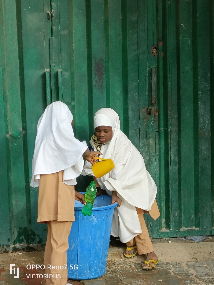 The image depicts two individuals, likely young females, engaged in an activity involving a blue plastic bucket against a dark green corrugated metal wall in Dutse, Nigeria. The individual on the left wears a white head covering and a tan long-sleeved garment, holding a partially visible green plastic bottle near the blue bucket. The individual on the right, wearing a white head covering, a white loose-fitting garment over tan trousers, and yellow flip-flops, is leaning forward and tilting a yellow plastic scoop, actively pouring a substance into the blue bucket. The blue bucket is ribbed and rests on an uneven, light-colored ground surface that shows wet patches. The background consists of a vertically corrugated dark green metal surface, possibly a door or wall, exhibiting rust, dirt, and a visible dark vertical handle with a locking mechanism. Both individuals are focused on the task involving the bucket.