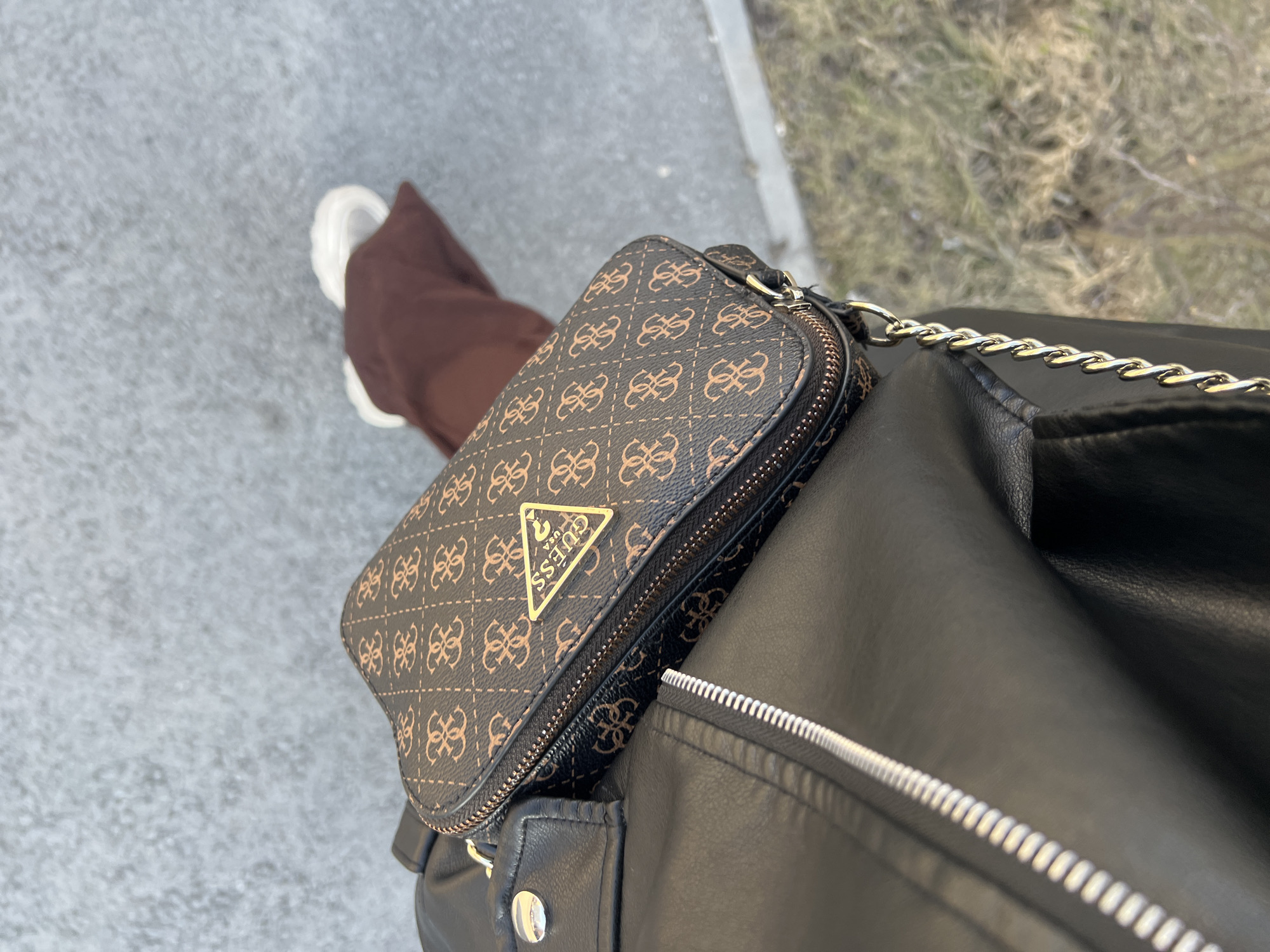 A first-person perspective shows a person wearing brown pants and white sneakers, carrying a black leather jacket with a silver zipper, and a brown Guess brand handbag with a gold logo. The person is walking on a gray pavement outdoors, possibly in the city of Tyumen, Russia, as the geotag indicates. The environment is slightly blurred, suggesting a focus on the clothing and accessories.
