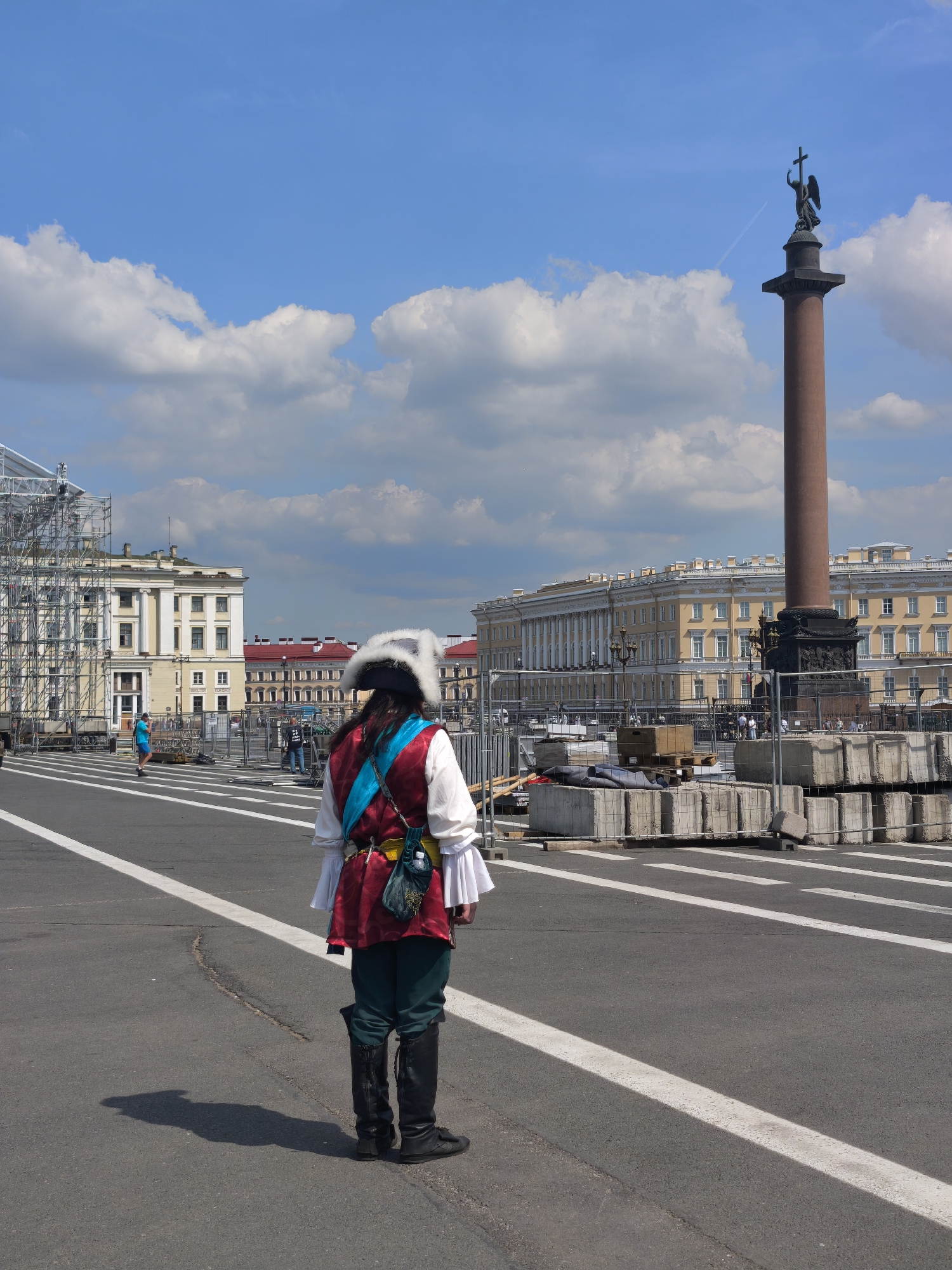 A person wearing a historical costume, including a white ruffled shirt, a red vest, teal pants, tall black boots, and a white fur-trimmed hat, stands in the foreground facing away from the camera. The person is positioned on an asphalt road with white lane markings. In the background, the Alexander Column, a prominent monument in Palace Square, Saint Petersburg, Russia, stands tall against a blue sky with scattered white clouds. To the left of the column, a large, light-colored building with classical architecture is visible, partially obscured by scaffolding. Construction barriers and stacks of concrete blocks are present in the middle ground, suggesting ongoing work or preparations for an event in the square. Other people, some in casual attire, can be seen in the distance near the buildings and the barriers. The overall scene depicts a sunny day in a public space in Saint Petersburg.