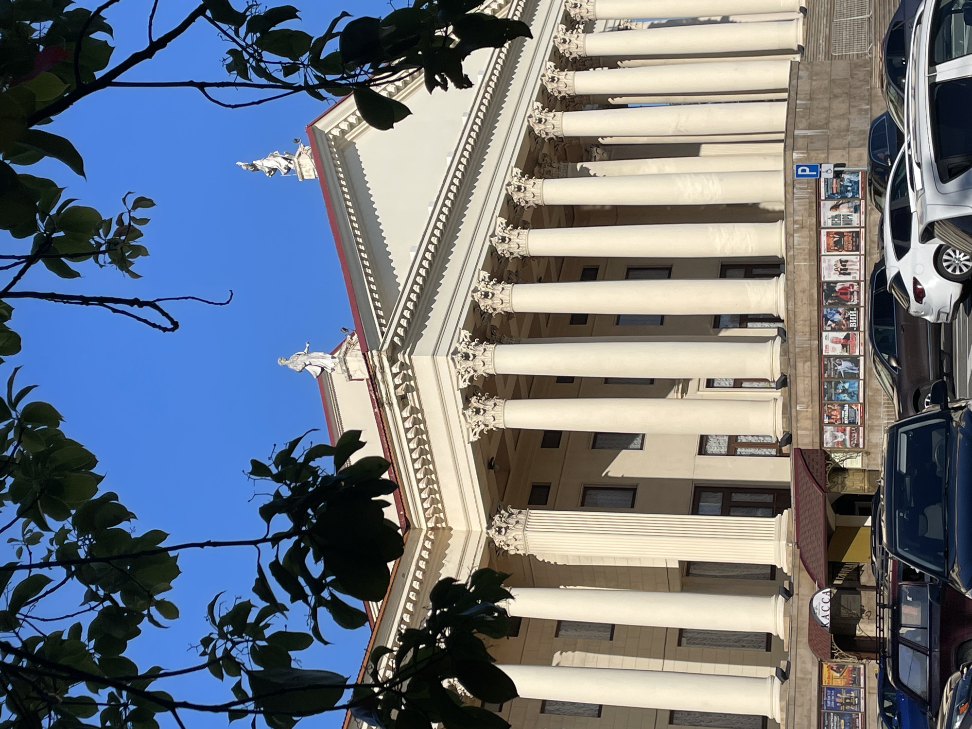 This is a photograph of the Winter Theater in Sochi, Russia, taken from across a street with cars in the foreground. The theater is a large, neoclassical building with a colonnade of tall, fluted columns supporting an entablature and a red-tiled roof. Two white statues are visible on the roofline. The facade is painted a pale yellow, and the columns are also a light color. Below the columns, at street level, there is a row of posters advertising performances. A sign reading "КАССА" (ticket office) is visible above an entrance. The sky is a clear, bright blue. Trees with dark green leaves partially obscure the top of the building. The street is paved, and several cars are parked or driving.
