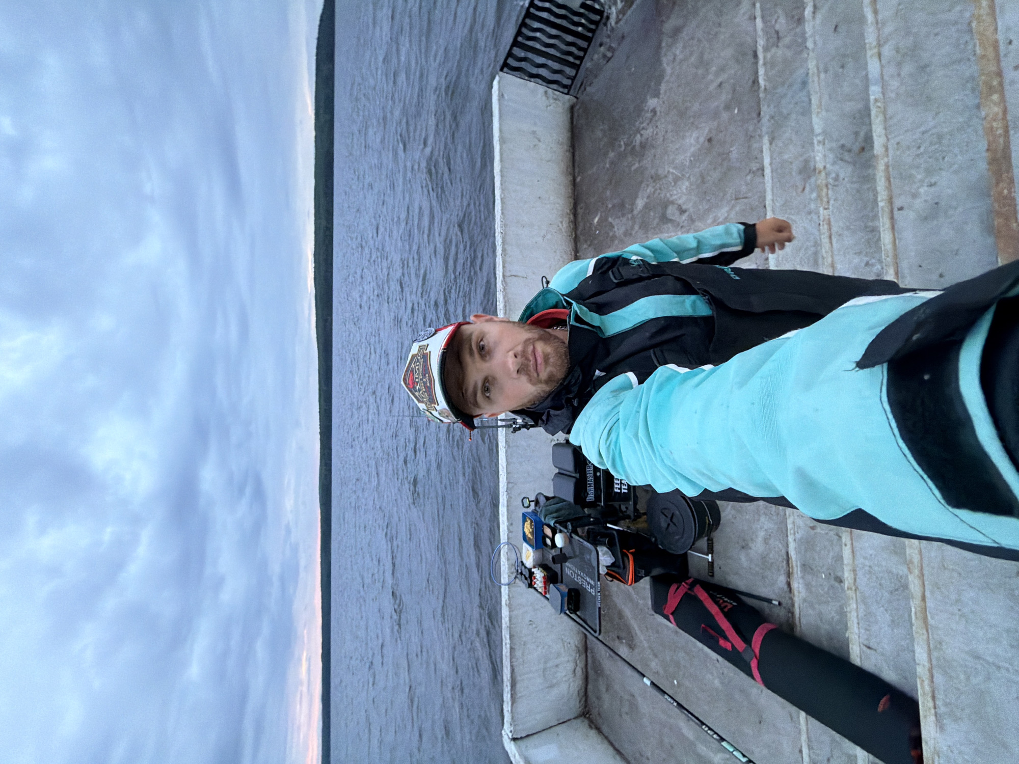 The image captures a male individual taking a selfie on a concrete pier or embankment overlooking a large body of water, likely a reservoir or lake, with a forested distant shore under an overcast sky. This setting is consistent with the natural environment surrounding Chaikovsky, Russia. The man, appearing to be in his 20s or 30s, has a beard and wears a multi-toned jacket (teal/light blue and black) over dark overalls, and a baseball cap with a prominent logo featuring the brand "MAXIMUS". His right arm is extended, holding the camera.
To the man's left, a fully equipped fishing station is set up on the concrete surface. This station includes a black seat box labeled "PRESTON INNOVATIONS" and "FEEDER TEAM", with an attached side tray holding multiple containers, a small blue tub containing food items, and a landing net. A fishing rod extends horizontally from the left side of the frame towards the water. Adjacent to the fishing station, a long black rod case with red straps and a black bucket are visible on the ground. The concrete platform features a low white wall separating it from the water. The body of water displays minor surface ripples and appears dark blue-grey. A continuous line of dark green trees forms the horizon, situated beneath a light grey, uniformly cloudy sky. A subtle light pinkish-orange hue is discernible at the very bottom edge of the sky, just above the distant treeline, suggesting either sunrise or sunset conditions.