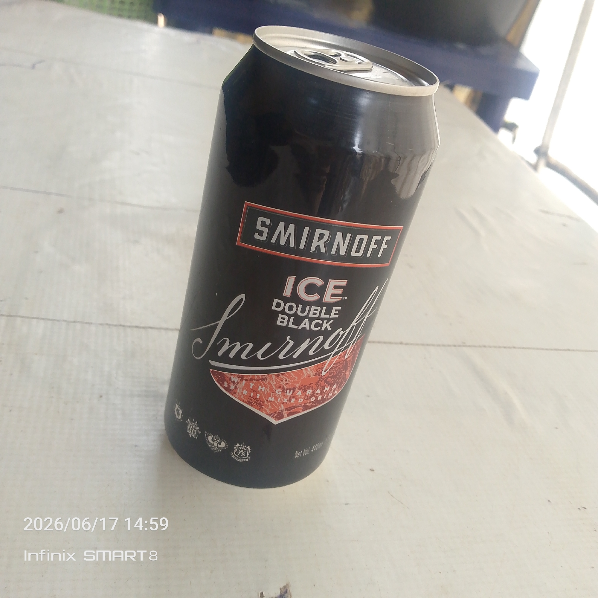 A close-up, slightly angled shot shows a black can of Smirnoff Ice Double Black. The can is the primary subject and is positioned in the center of the frame, tilted to the right. The background is a light-colored surface, possibly a table or countertop, with some faint lines suggesting texture or a pattern. The lighting appears to be natural, casting soft shadows and highlighting the metallic sheen of the can.
The label on the can is clearly visible. At the top, in a red-bordered rectangular box, is the word "SMIRNOFF" in white capital letters. Below this, in a larger, stylized font, is "ICE". Further down, in smaller white text, it reads "DOUBLE BLACK". A decorative script with the word "Smirnoff" is emblazoned across the lower portion of the label, with a red, textured graphic underneath. Small icons and text are visible near the bottom of the can, including "Vet Vol. 440m". In the bottom left corner of the image, there is text that reads "2026/06/17 14:59 Infinix SMART 8", indicating the date and time the photo was taken, as well as the device used.
The overall mood of the image is neutral, focusing on the product itself. There are no people or specific environmental cues that suggest a particular location or weather conditions. The scene is simple and uncluttered, drawing attention solely to the Smirnoff can.