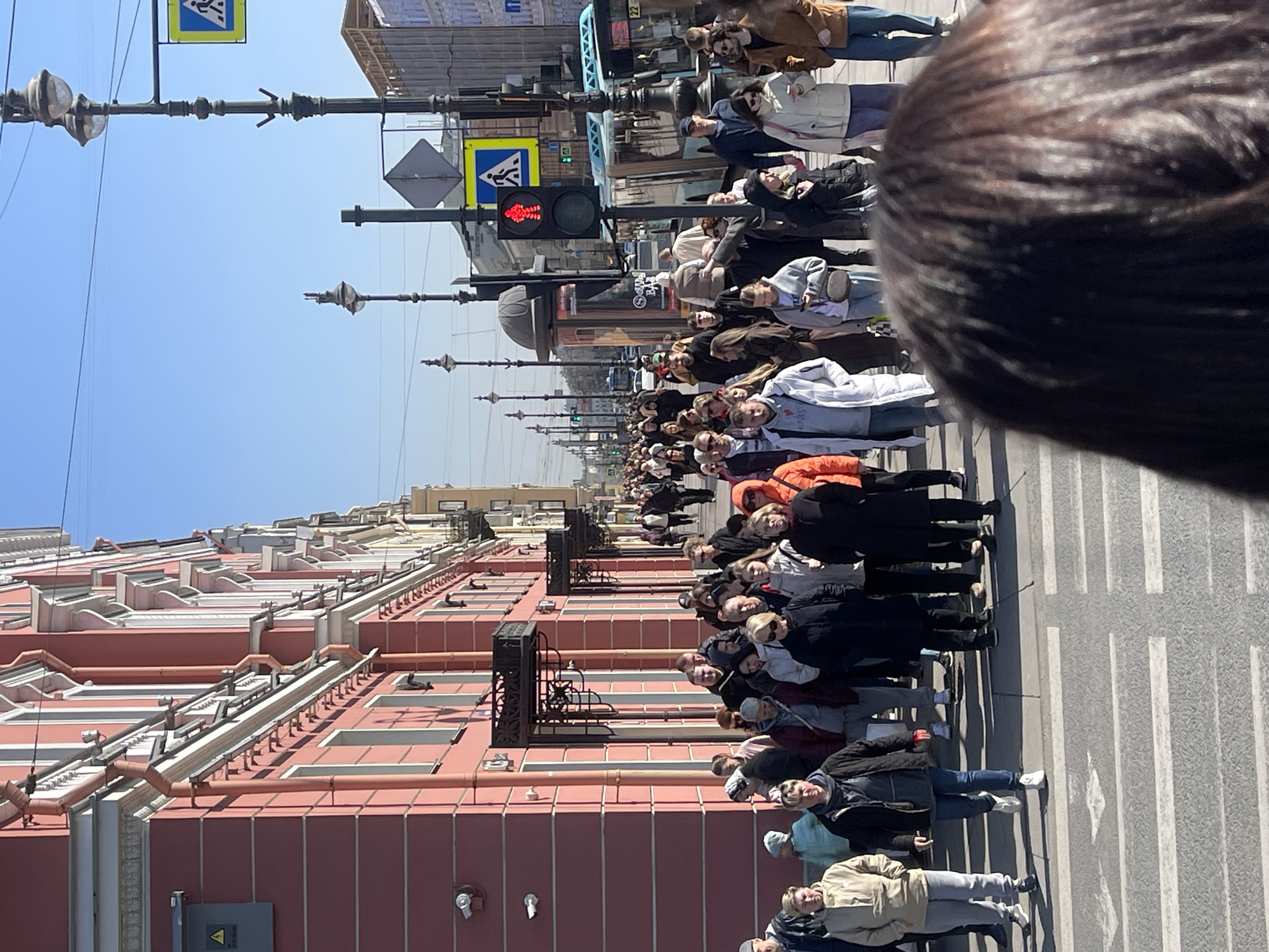 The image captures a street scene in Saint Petersburg, Russia. A large crowd of people, predominantly adults, are standing on a crosswalk. The pedestrian crossing signal shows a red light. Most of the people are facing the camera. They are dressed in a variety of casual clothing, including jackets and sweaters.
On the left side of the frame, there is a pink building with multiple windows and some metal fixtures. On the right side, there are street lights and traffic signals with a pedestrian crossing sign. The sky is clear and blue, indicating a sunny day. The perspective suggests the photo was taken from a high vantage point. The top right of the image features the back of a person's head.