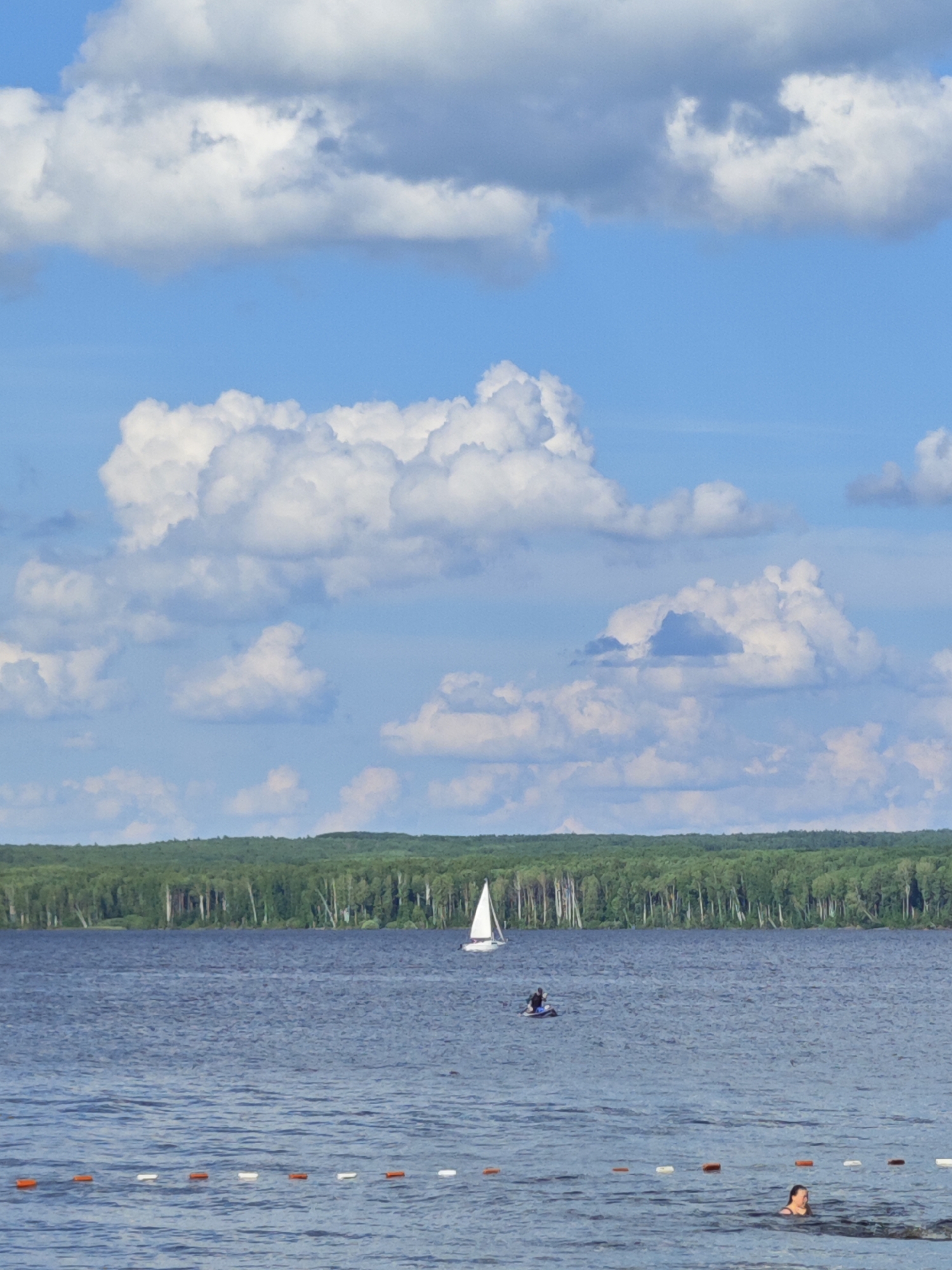 This media file captures a serene summer day at a lake, likely in Chaikovsky, Russia. The scene is dominated by a vast expanse of calm, blue water, reflecting the bright sky above. The sky itself is a canvas of vibrant blue, dotted with fluffy, white cumulus clouds that suggest fair weather.
In the distance, a lush green forest line forms a dense backdrop to the lake, with tall trees creating a natural horizon. The time of day appears to be midday or early afternoon, given the bright sunlight and the shadows cast by the clouds.
The water is populated with a few individuals engaging in leisurely activities. A small sailboat with a white sail is gracefully gliding across the water, adding a touch of tranquility to the scene. Closer to the shore, a person is seen on a paddleboard, enjoying the calm water. Further in the foreground, another person is swimming, their head visible above the surface, suggesting a refreshing dip on a warm day.
A rope with buoys, some orange and some white, stretches across the water in the foreground, likely marking a designated swimming area or a boundary. The overall atmosphere is one of peaceful recreation and enjoyment of nature on a pleasant summer day. There is no visible text in the image.