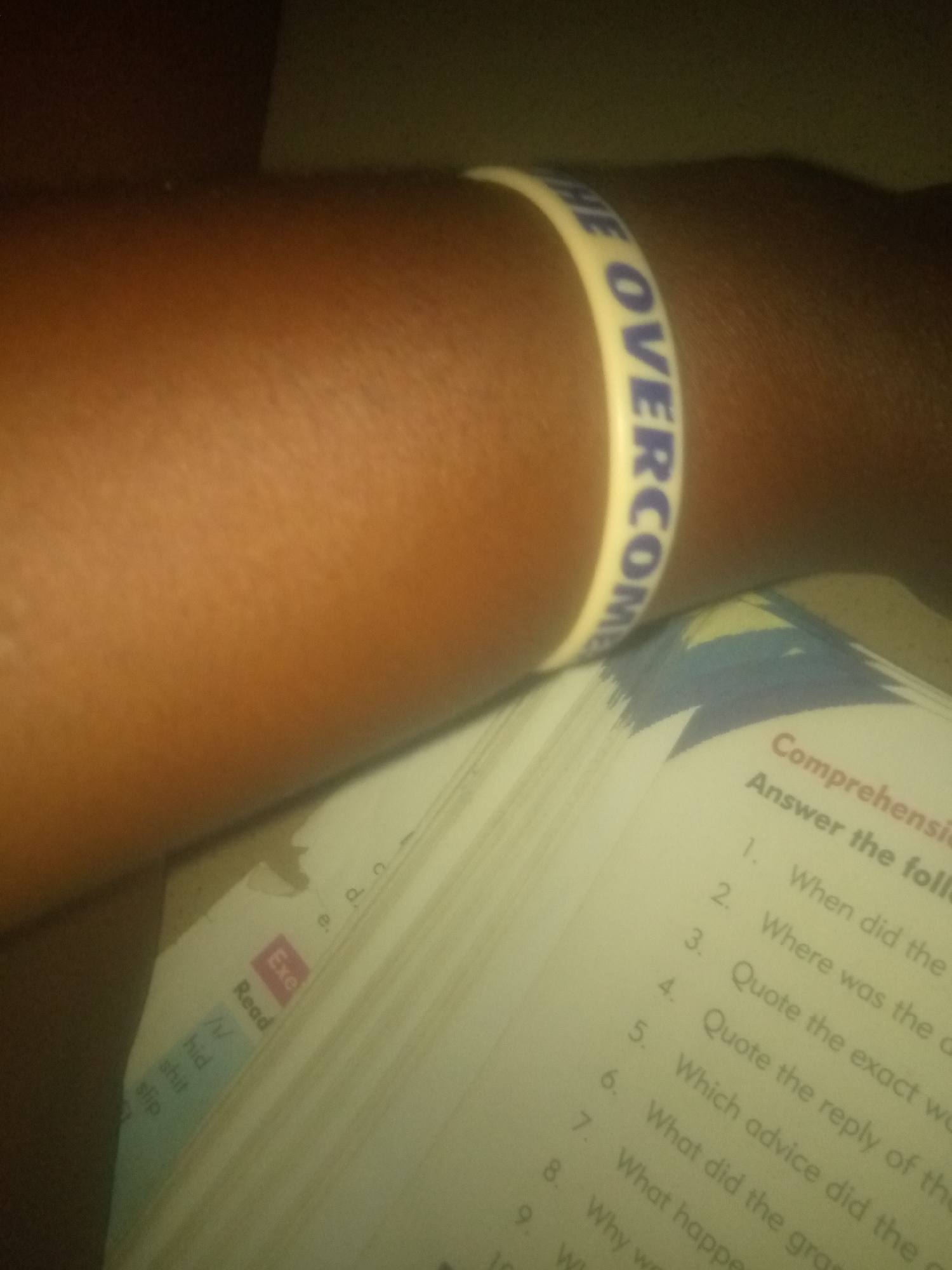 The image shows a close-up view of a person's arm wearing a white wristband with blue lettering that reads "THE OVERCOME". Part of an open book with questions is visible in the foreground. The book has a title that appears to be "Comprehensions" and numbered questions such as "1. When did the", "2. Where was the", and "3. Quote the". There are also some words on a tab on the left side of the book, including "Exe" and "Read". The background is dark and out of focus. The image is likely taken indoors, possibly in a home or classroom setting. There is no visible indication of the specific location within Jauro Yinu, Nigeria.