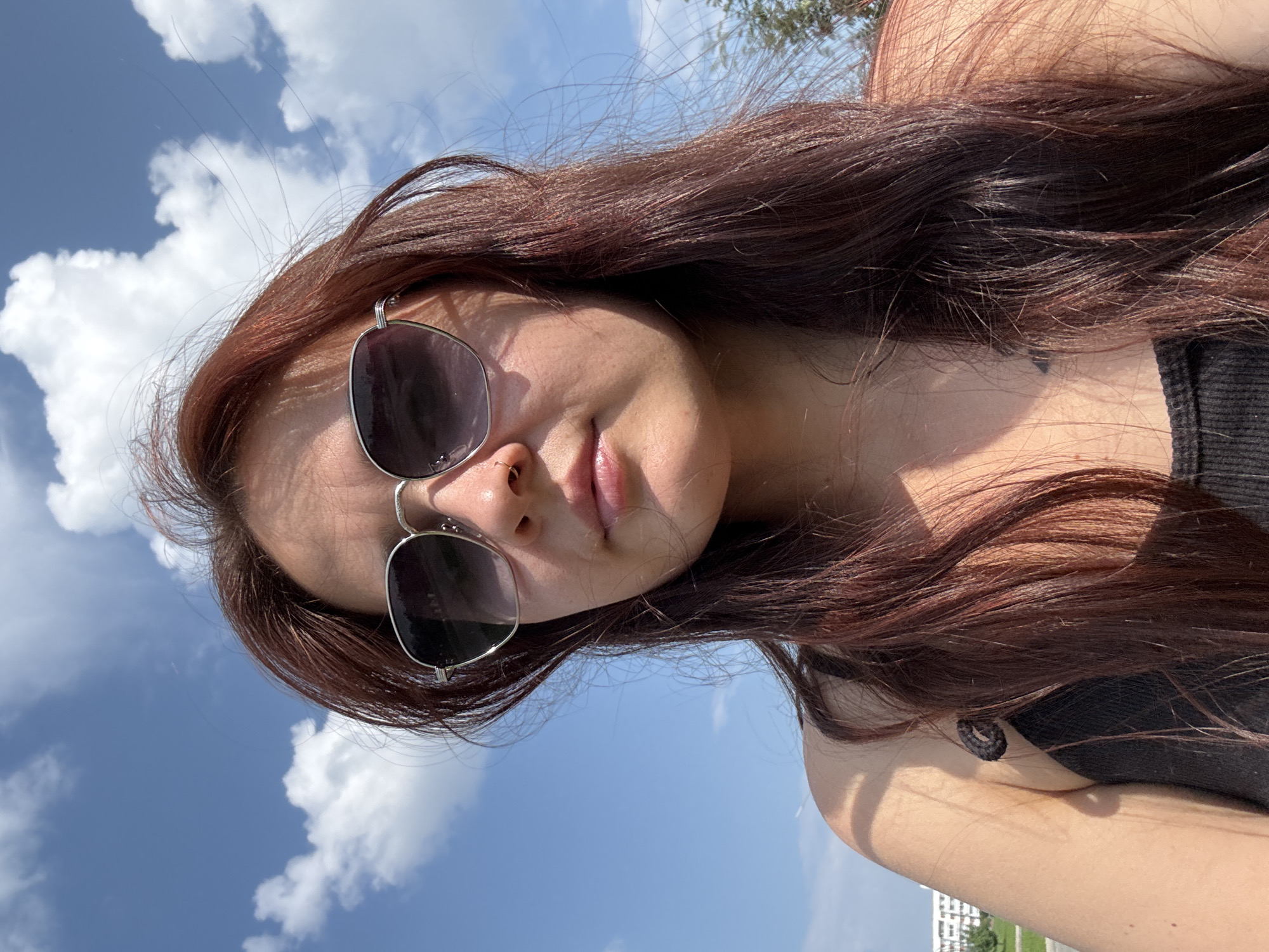 The image shows a close-up selfie of a woman with long, dark red hair wearing sunglasses and a black tank top. She has a nose ring and is outdoors with a bright blue sky and white clouds in the background. The setting appears to be a city, with buildings visible in the distance on the left side of the frame. There are trees on the right side of the frame. The lighting suggests it is daytime, likely sunny. No specific landmarks are identifiable to pinpoint a location within Naberezhnye Chelny, Russia.