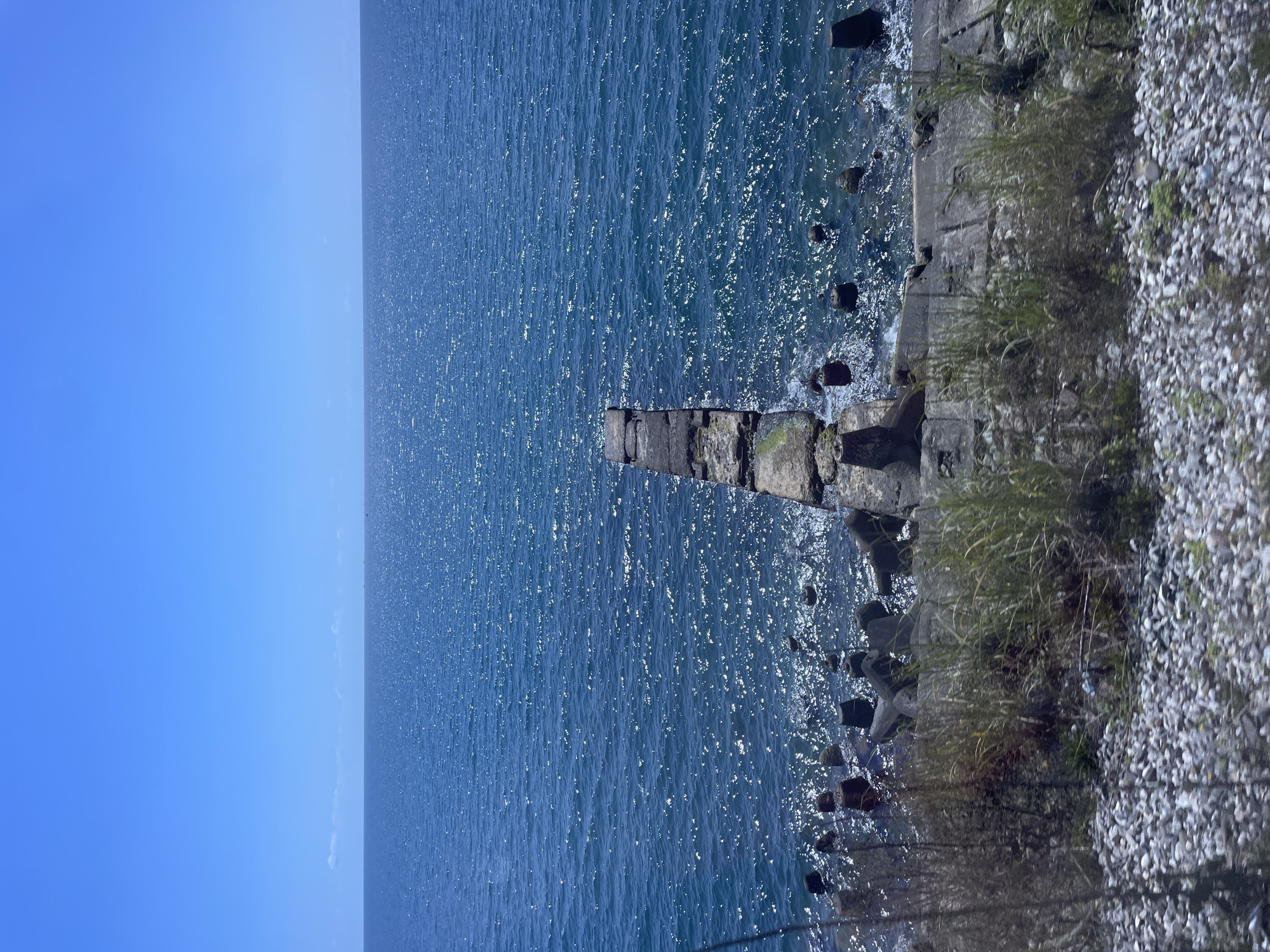 A clear, bright, sunny day is depicted with a vast expanse of deep blue ocean stretching to the horizon under a cloudless, pale blue sky. In the foreground, a weathered concrete pier extends out into the water, its surface uneven and showing signs of age. The water is a shimmering expanse of blue, with sunlight glinting off its surface, creating a textured, sparkling effect. Along the shoreline, a rocky embankment is visible, interspersed with concrete structures and vegetation. The scene appears to be a coastal area, possibly a breakwater or a disused pier. While the exact location within Sochi, Russia, cannot be definitively identified from the image alone, it captures a serene and natural coastal environment. There are no people or animals visible in the image.