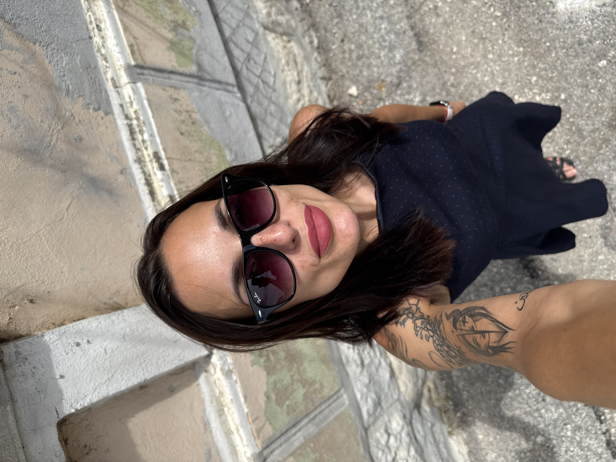 A woman in a dark blue polka dot dress and sunglasses is taking a selfie outdoors in Pleven, Bulgaria. She is standing near a weathered stone wall and a paved area. Her left arm is visible, displaying multiple tattoos.