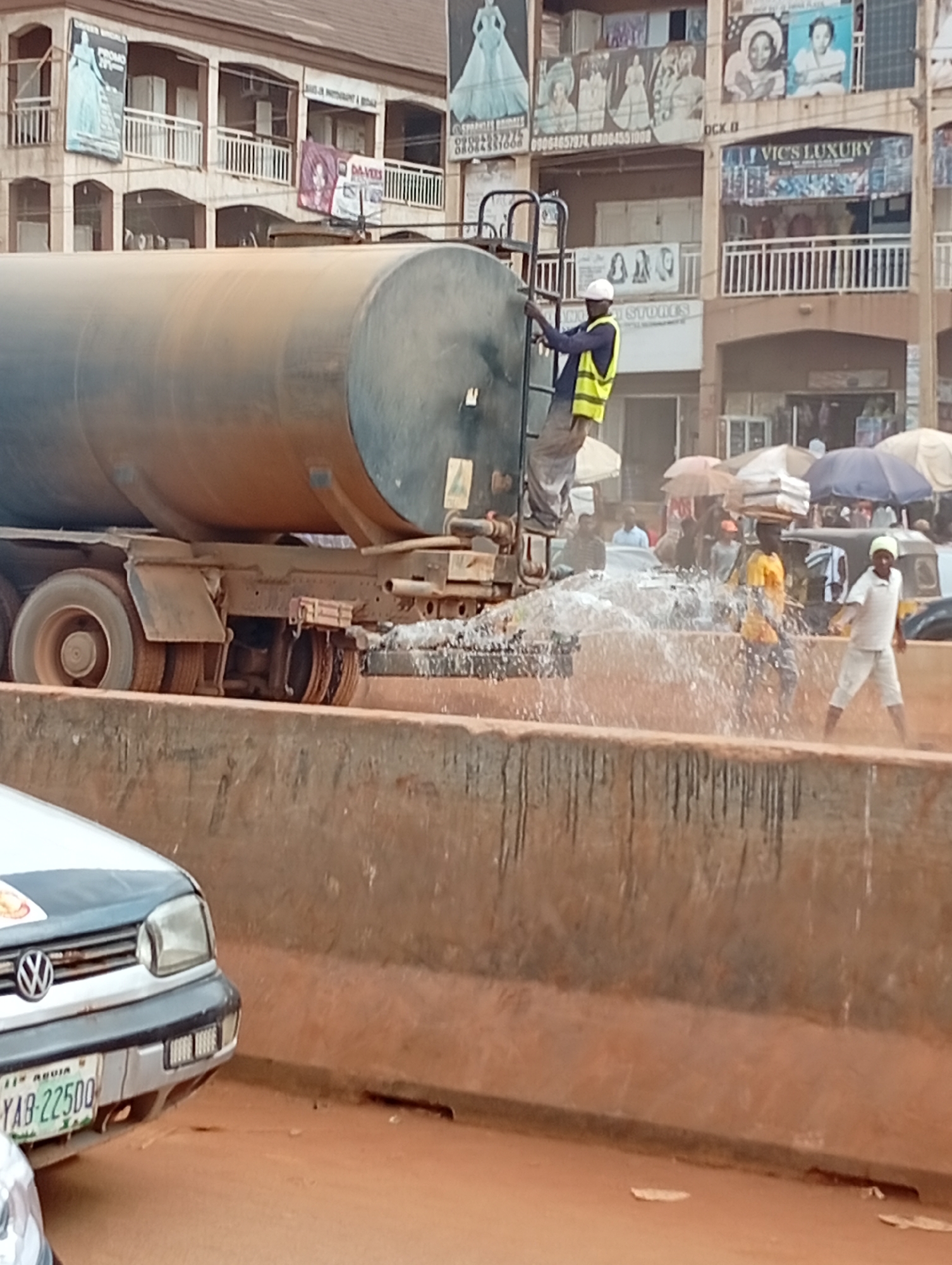 The scene depicts a water truck watering a street in Ado, Nigeria. A worker in a white hard hat and a yellow safety vest is standing on a ladder attached to the side of the large tank truck, seemingly operating the water release mechanism. Water is spraying out of the truck, creating a mist over the street and some people walking nearby. One person is carrying objects on his head. A car is visible on the left of the frame with a license plate that reads "YAB-22500". The background includes multi-story buildings with storefronts and advertisements on the upper levels.