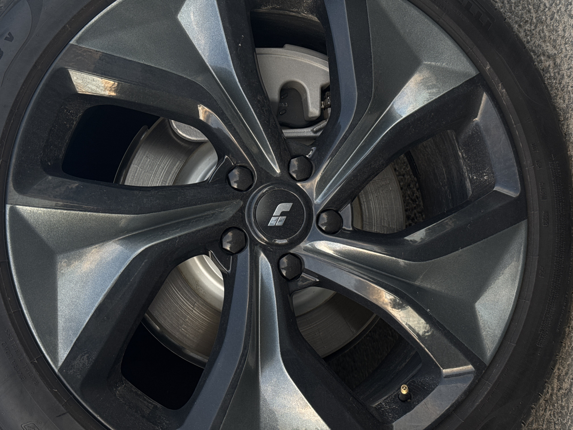 The image presents a close-up, eye-level view of a car wheel and tire, likely from a vehicle parked on a textured, possibly paved, surface. The multi-spoke wheel is predominantly dark gray with an intricate design, featuring alternating matte and reflective, possibly machined or polished, surfaces on its seven main spokes.
In the center of the wheel, a black hub cap displays a silver, stylized logo resembling a geometric "C" or an abstract symbol. Five black lug nuts are arranged around the hub cap. Through the spokes, the vehicle's braking system is visible. A silver-colored brake caliper is positioned behind the upper spokes, with "315.3" and other partially legible alphanumeric markings visible on its surface. The metallic brake rotor behind the caliper shows concentric wear patterns from use.
The black tire mounted on the wheel displays various sidewall markings. Visible text includes "PIRELLI" (partially), "RADIAL TUBELESS", "EXTRA LOAD", "M+S" (indicating Mud+Snow), "DOT 06X", and other codes such as "245/45 R20" (implied tire size) and "103W" (load and speed rating). A brass-colored valve stem cap is present on the lower part of the wheel. The bright lighting suggests it is daytime, with strong highlights reflecting off the wheel's surface. The wheel shows some light dust or residue.
