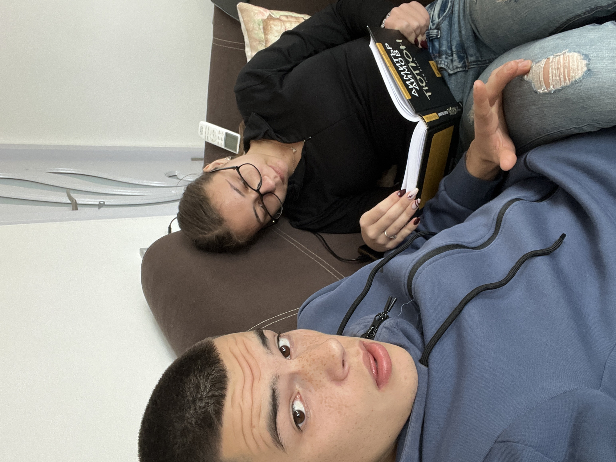 Two people are lying on a brown sofa. The person in the foreground, likely male, is wearing a blue hoodie and has short dark hair and freckles. They are looking directly at the camera with a slightly surprised expression. Behind them, a person wearing a black hoodie and glasses is lying down, reading a book titled "АНДРЕЙ ДМИТРЕВ АКМГБР." This person is wearing jeans with a hole in the knee. A remote control is visible on the sofa near the reader. The background is a white wall with a decorative silver design. The location is not discernible from the image and cannot be reliably determined as Karavelovo, Bulgaria.