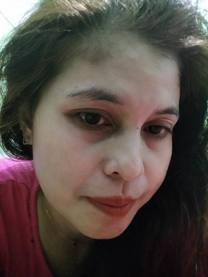 A close-up, head-and-shoulders portrait of an individual, likely female, is depicted, taken in the city of Ranugedang, Indonesia. The subject possesses dark brown, wavy hair framing a fair-skinned face. Distinct dark eyebrows arch above dark eyes, which show evidence of dark circles beneath them. A small dark mark is present above the left nostril. The lips are colored with a reddish-brown lipstick. The subject wears a pink garment, visible on the left, and a thin gold-colored necklace with a small, dark, quatrefoil-shaped charm. A small gold-colored hoop earring is present in the left ear. The subject's head is slightly tilted, and the gaze is directed downwards and to the left. The background is blurred, indicating a shallow depth of field, with soft, ambient lighting present. No direct interaction is observed.