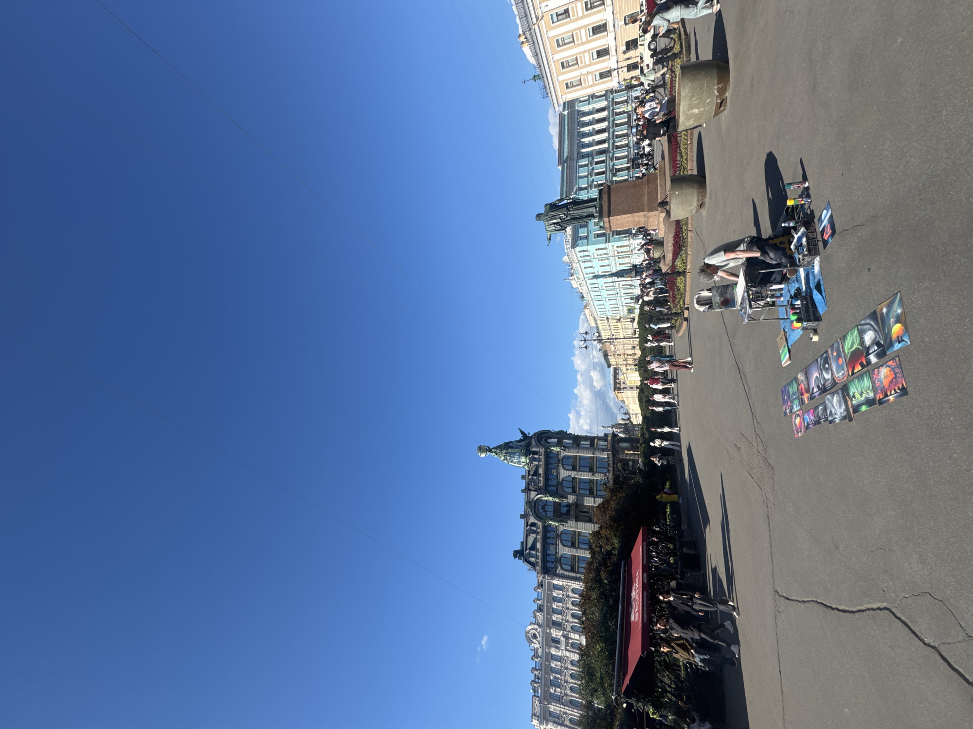 A street scene in Saint Petersburg, Russia, features a clear blue sky with a few wispy clouds. The image is oriented vertically, making it appear as if the viewer is looking up at a cityscape. In the foreground, a man is seated near a wheeled cart filled with art supplies, including paint cans and brushes. Several paintings, depicting abstract and cosmic scenes, are displayed on the ground in front of the cart and also further down the sidewalk.
Beyond the artist and his display, a crowd of people walks along a paved walkway. The walkway is bordered by a low red awning and trees. In the background, grand European-style buildings with ornate architecture line the street. A notable statue of a man on horseback is visible in the mid-ground, positioned in front of a light blue building. Further down the street, another impressive building with a green domed roof is prominent. The sunlight suggests it is daytime, likely midday, given the bright sky and shadows cast by the people and objects. The overall atmosphere appears calm and leisurely, with people enjoying a sunny day outdoors and an artist showcasing their work.