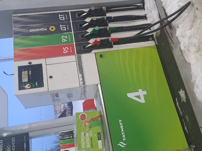 The image captures a gas pump at a fuel station, likely during daytime in cold weather, indicated by patches of snow on the ground. The photograph is taken at an oblique angle, making the pump appear tilted.
The central focus is a modern, light-colored fuel dispenser unit. It features multiple fuel nozzles hanging on its right side, each connected to a hose. From top to bottom, these appear to be a black nozzle, a green nozzle, and a red nozzle. Corresponding colored panels on the pump body indicate the fuel types: a black panel for "ДТ" (DT, likely diesel fuel), a green panel for "92" (likely 92 octane gasoline), and a red panel for "95" (likely 95 octane gasoline). Another black panel with "ДТ" is visible further up, next to "ТОНЕКО" (TOHEKO), suggesting another diesel option or branding.
On the left side of the pump, there is a digital display panel. The display shows two sets of numbers, "139.76" and "20.00", which are typically indicative of the total cost and the volume of fuel dispensed (e.g., 20 liters for 139.76 units of currency). Below the display, there is a small green button or indicator.
To the right of the fuel dispenser, a large, predominantly green panel is visible, displaying a prominent white number "4". Below this number, the "TATNEFT" logo is clearly visible, consisting of a stylized flame icon and the company name. Another "TATNEFT" logo is partially visible at the bottom left of the image.
Below the main pump unit and partially obscured, another green panel or sign displays Cyrillic text: "ЧЕМПИОНЫ ЭКОЛОГИИ" (Champions of Ecology). Further down, partially visible text includes "НЕКО" (NECO) and "ЛОНОВ!" (part of a word, possibly "МИЛЛИОНОВ!" - millions).
The background shows a clear blue sky, a street light pole, and some blurred structures that might be parts of buildings or trees. Snow is accumulated along the bottom edge of the pump unit and on the ground.