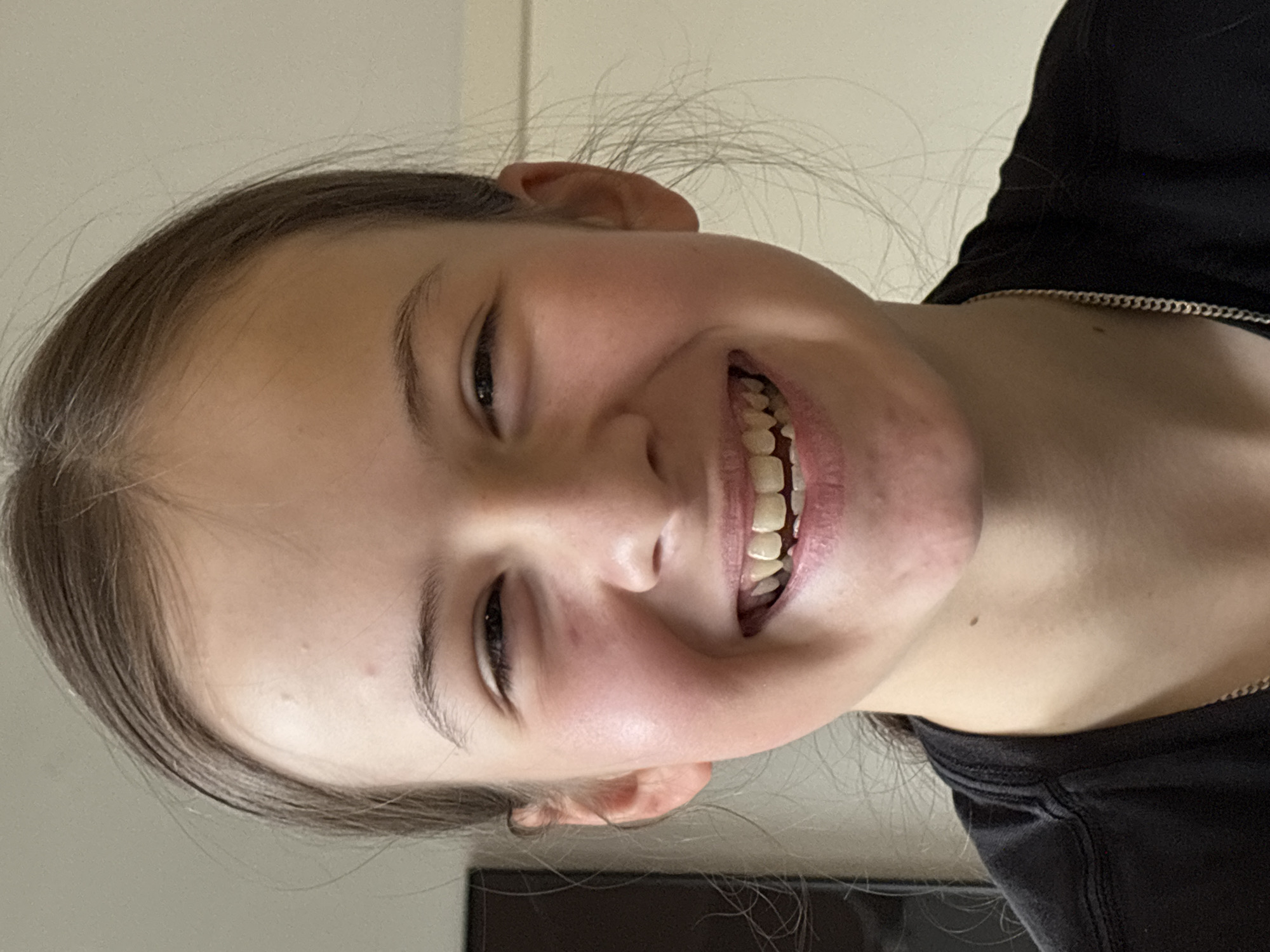 The image displays a close-up portrait of a young person, likely female, with light brown hair and fair skin, captured in an indoor setting against a plain, light-colored background. The individual is smiling broadly, showing both upper and lower teeth, which convey a sense of happiness and cheerfulness. Her dark eyes are open and slightly crinkled at the corners from the smile.
Her hair is mostly pulled back from her face, with some fine, wispy strands visible around her forehead and ears. She is wearing a dark, possibly black, crew-neck top, and a thin, metallic chain necklace is visible around her neck. The image is rotated counter-clockwise, making the subject's head appear significantly tilted to the right, with her gaze directed slightly upwards. No specific time of day or weather can be determined from the visual information. There are no distinct location cues within the image itself, other than an ambiguous indoor environment. No text is visible in the media file.