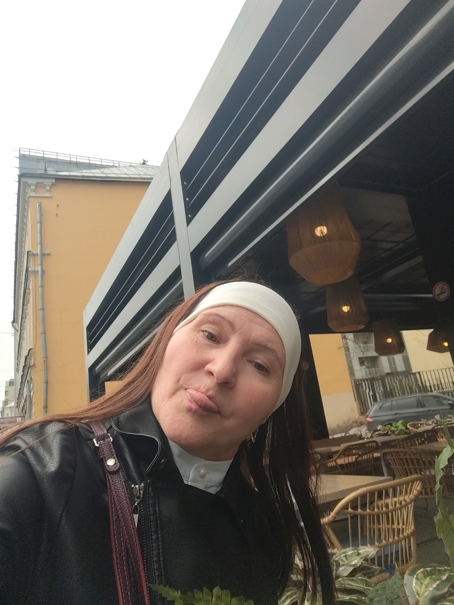 A woman with reddish-brown hair and a white headband takes a selfie outdoors, looking directly at the camera with a playful expression, her lips slightly puckered. She is wearing a black leather jacket over a light-colored collared shirt, and a dark red shoulder bag strap is visible.
The scene appears to be an urban outdoor dining area in Moscow, Russia. Behind her on the left is a tall, light yellow building with traditional architectural details and a grey drainpipe, set against an overcast sky. To her right, an awning or part of a building features distinctive black and white horizontal stripes. Underneath this structure, several wicker-style pendant light fixtures emit a warm glow.
Further into the background on the right, there are numerous outdoor cafe tables and chairs made of woven material, along with potted plants, some with variegated leaves. A dark-colored station wagon or SUV is parked on a street behind the cafe furniture. A circular "No Smoking" sign, depicting a crossed-out cigarette, is visible on a pillar near the hanging lights. The overall lighting suggests daylight on an overcast day.