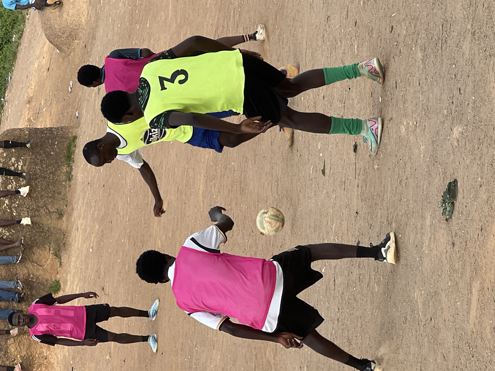 An outdoor, ground-level, full-shot photograph captures a group of young males playing soccer on a dirt field. The scene appears to be in Jos, Nigeria. In the foreground, a young male wearing a pink vest over a white and black shirt and black shorts is positioned on the left, facing away from the viewer with his left leg extended towards the soccer ball in mid-air. To his right, a young male in a green vest with the number '3' on the back, black shorts, and green socks is also facing away from the viewer, with his right leg extended. Another young male in a yellow vest over a white t-shirt and blue shorts is visible in the middle ground, facing left and partially obscured. In the background, other individuals are seated on a raised, rocky, and uneven surface. The ground is predominantly dusty brown, with scattered debris. The lighting suggests it is daytime.