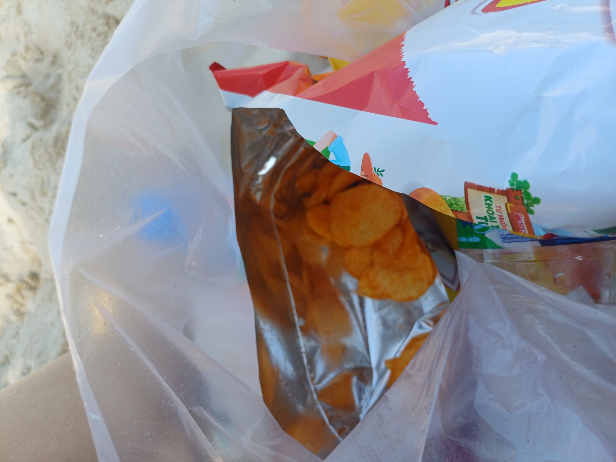 This close-up image shows a plastic bag filled with potato chips, situated on sand, suggesting a beach setting. The bag is mostly translucent, with some chips visible inside through the plastic. One partially opened bag of potato chips, with a red and white design, is prominent within the larger bag. The chips appear to be of a standard potato chip variety, with an orange hue. The background is composed of light-colored sand, characteristic of a beach in Nha Trang, Vietnam. A person's leg, clad in light-colored fabric, is partially visible in the upper left corner. The scene is brightly lit, indicative of daylight.