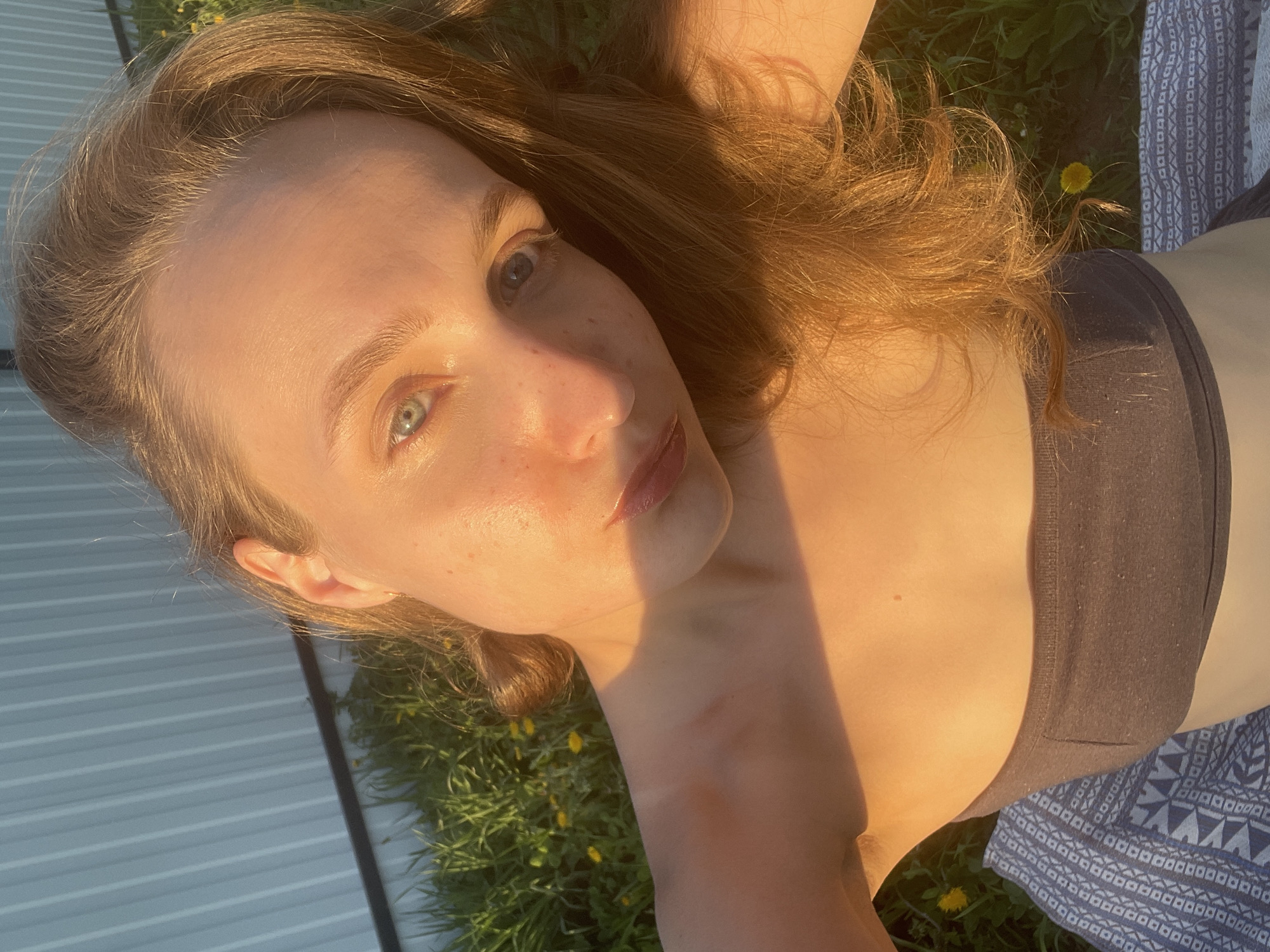A woman is lying on the grass, possibly in Подшивалово, Russia. She is wearing a dark top and patterned pants. The sun is shining brightly, creating strong shadows. There are dandelions in the grass. She is lying near a metal structure, which is possibly part of a building.