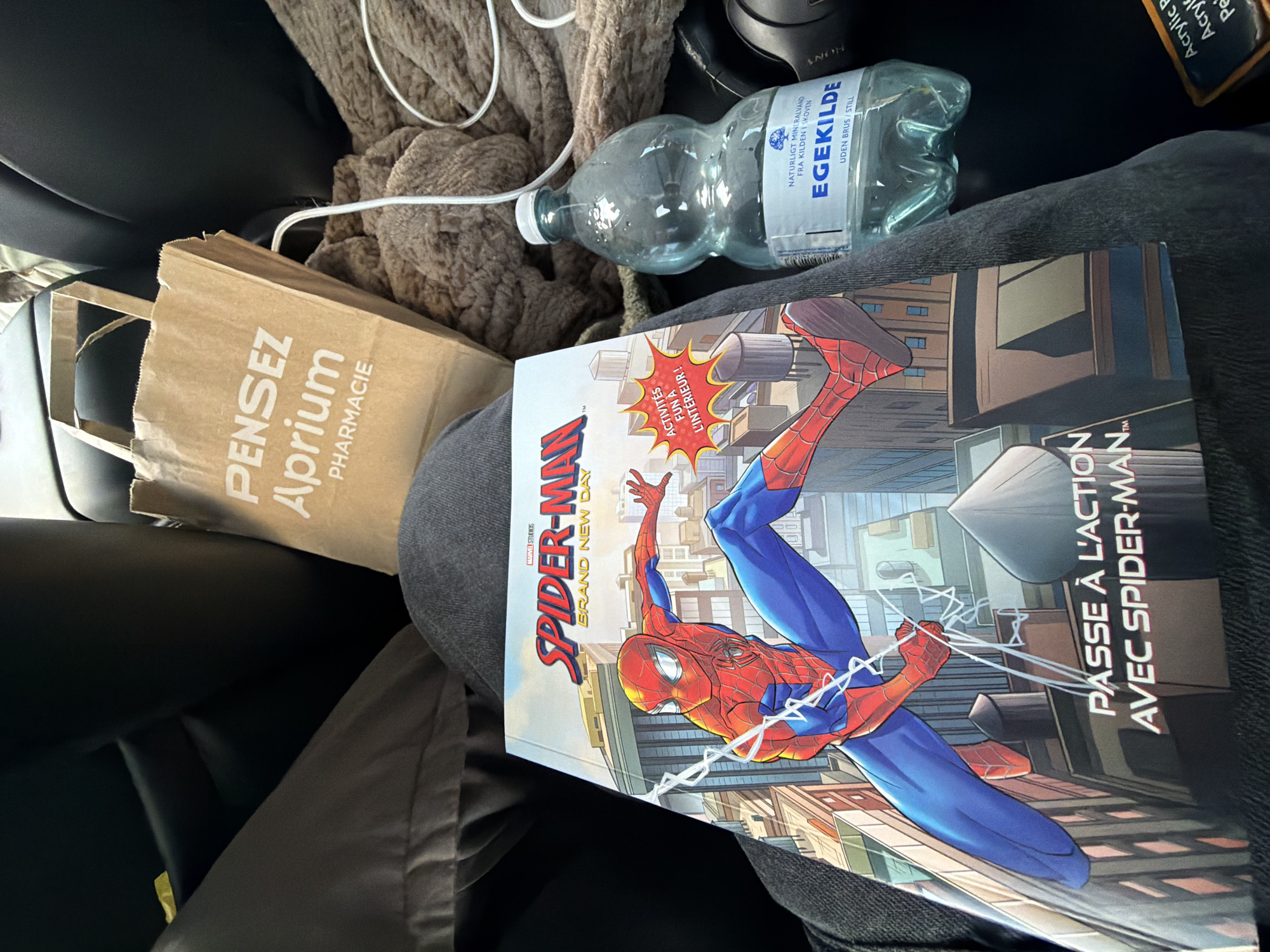 A close-up shot captures a person's lap in a car interior, with various items resting on it. The primary subject is a Spiderman comic book, featuring the character in a dynamic pose against a cityscape. To the right of the comic is a brown paper bag with white text that reads "PENSEZ Aprium PHARMACIE". Further to the right, a clear plastic bottle of EGEKILDE natural mineral water sits prominently, its label indicating it is from a source in the forest. A brown, textured blanket is draped behind the water bottle. The background shows the dark interior of a car, with the edge of a seat visible. The image appears to have been taken from a seated perspective within the vehicle, suggesting the person is either a passenger or the driver. The scene does not provide any specific visual cues to determine the location within the city of Ödsmål, Sweden, as there are no identifying landmarks or signs visible.
