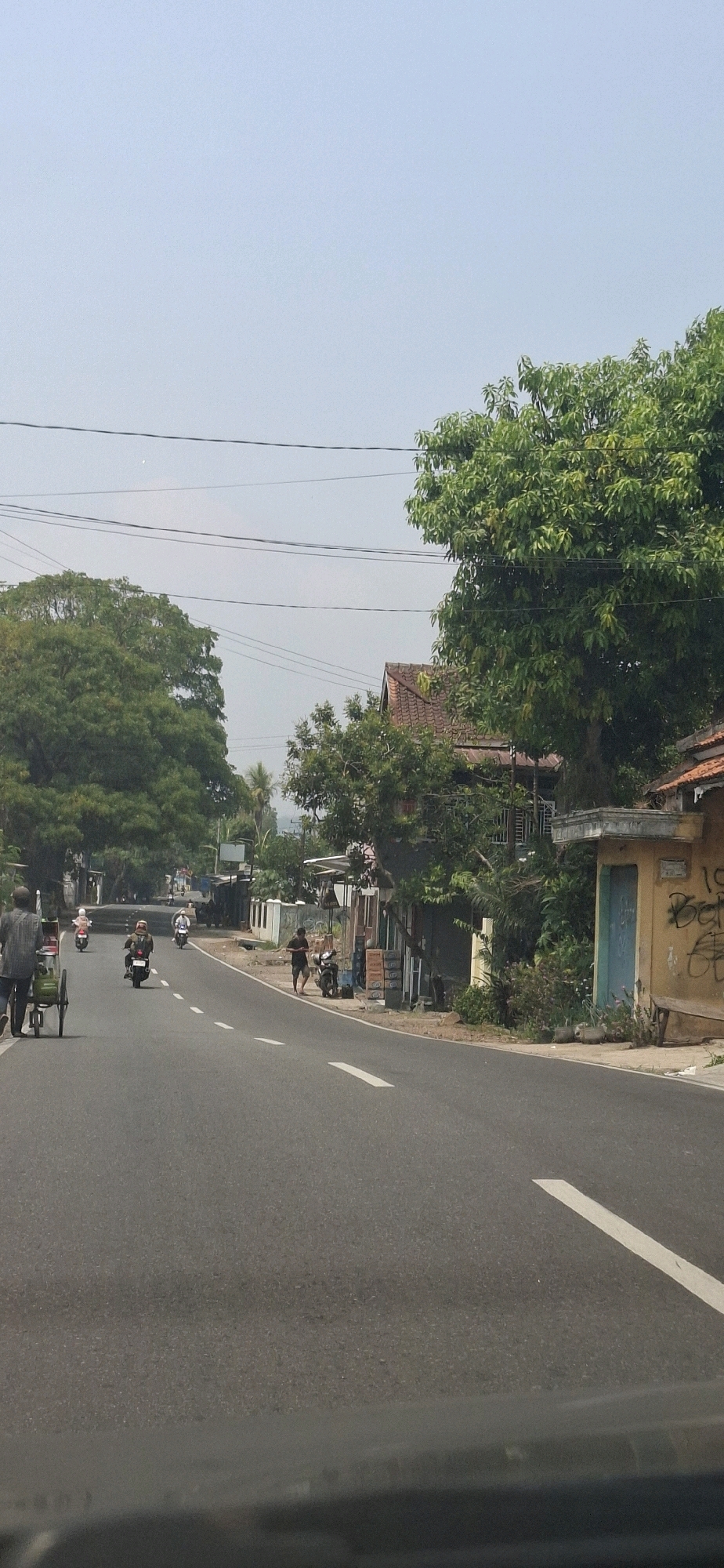 This is a vertical, eye-level, medium shot photograph taken on a sunny day in Susukan, Indonesia. A two-lane asphalt road dominates the foreground, with a white broken line marking the center and a solid white line on the right shoulder. On the left side of the road, a man in a checkered shirt walks next to a green handcart. Further down the road, several people on motorcycles are visible.
The right side of the road features a row of low-rise buildings with various storefronts and awnings. One building, painted a pale yellow, has graffiti on its facade and a blue door. A wooden bench is placed in front of this building. Dense green foliage, including large trees, lines the street and the buildings, with some trees reaching towards the sky and obscuring the horizon. The sky is clear and light blue. Electrical wires are strung across the upper portion of the image. The overall impression is of a somewhat rural or suburban street scene.