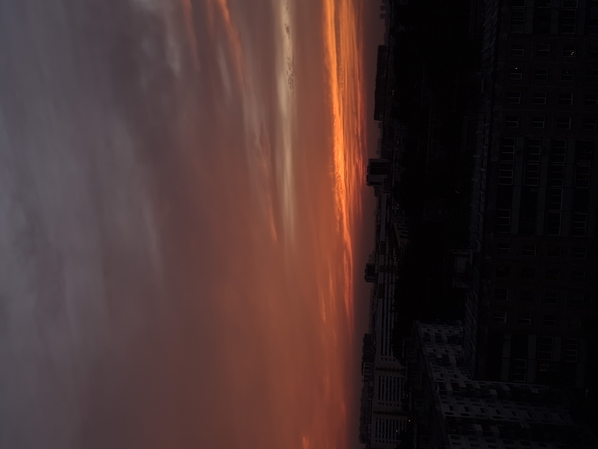 This image depicts a dramatic sunset over a cityscape, likely Novosibirsk, Russia, given the architecture. The sky is ablaze with fiery hues of orange, red, and pink, transitioning into muted grays and purples towards the top of the frame. Streaks of clouds catch the fading light, creating a textured and dynamic celestial display. Below the vibrant sky, a silhouetted urban landscape unfolds. The foreground is dominated by dark, shadowy apartment buildings, their forms partially obscuring the city's middle ground. The architectural style suggests Soviet-era housing blocks, characterized by their uniform facades and numerous windows. In the distance, more buildings and what appear to be trees are visible, also rendered in silhouette against the luminous sky. The overall impression is one of urban twilight, with the natural spectacle of the sunset providing a stark contrast to the man-made structures below. No people are visible in the image.