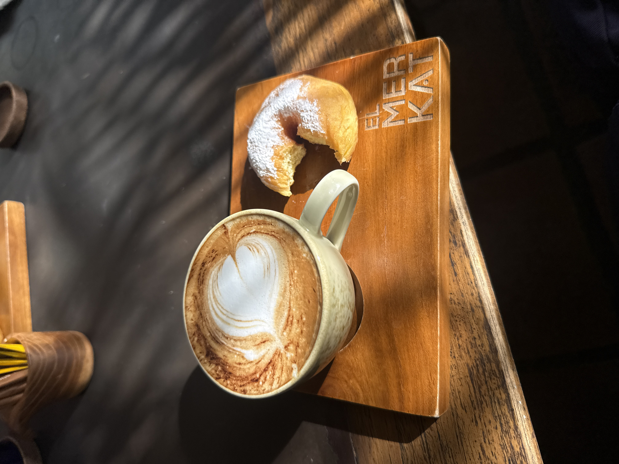 The image shows a cappuccino and a doughnut on a small wooden serving board, labeled "EL MER KAT", likely a reference to El Merkat restaurant in Kuta Selatan, Indonesia. The doughnut is dusted with powdered sugar and appears to have a bite taken out of it. The cappuccino has latte art with a heart shape. The scene is bathed in bright sunlight, possibly outdoors, with shadows cast across the table. The table itself is made of dark wood. A toothpick holder and some items with circular tops are also visible on the table.