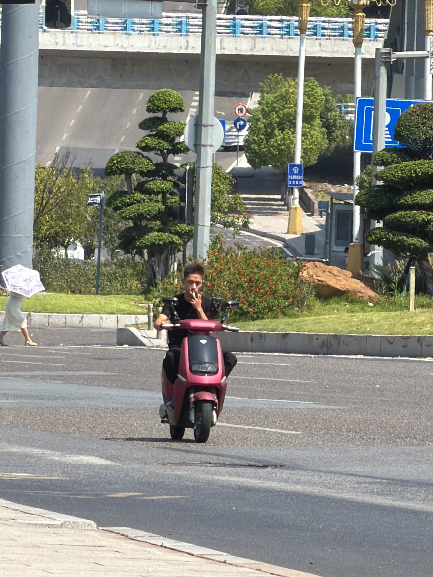 In a sunlit urban scene in Jiangjin, China, a young man on a pink electric scooter is captured mid-action, smoking a cigarette. He is wearing a dark t-shirt and is facing forward, his right hand on the scooter's handlebar. The scooter has a red body and a black seat, with its front wheel visible. To the left of the scooter, a woman in a light-colored dress walks with a white, translucent umbrella. The background features a road with gray asphalt and white lane markings. Beyond the road, a green grassy area is visible, with several stylized trees, one of which is a prominent topiary. In the far background, a concrete overpass or bridge with blue railings is visible. The overall environment appears to be a well-maintained urban roadside.