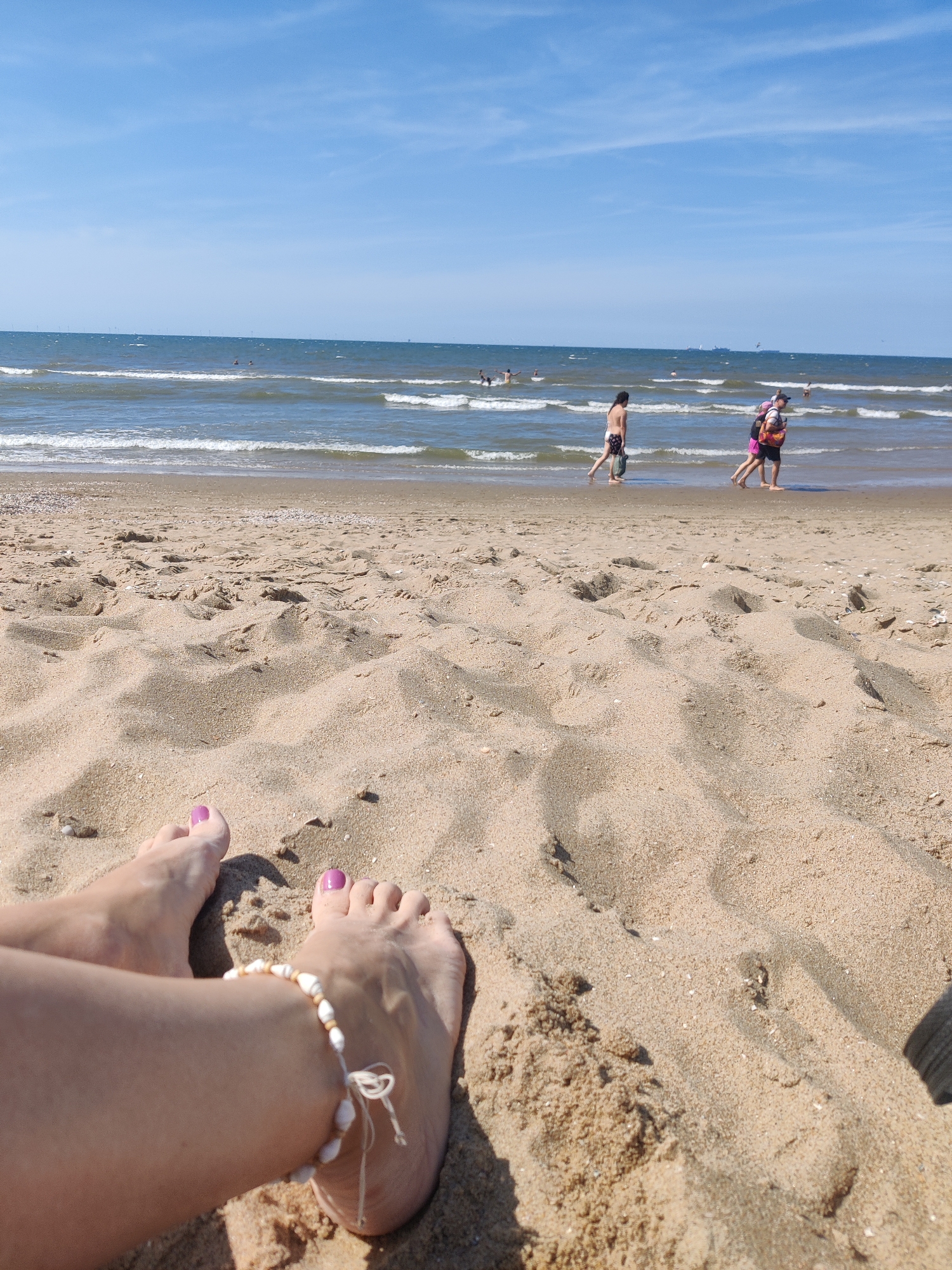 A low-angle, close-up shot shows a person's feet resting on a sandy beach. The toes are painted with purple nail polish, and a shell anklet adorns one ankle. The sand is textured with ripples and small shells. In the background, the ocean meets the horizon under a bright blue sky with scattered white clouds. Several people are visible in the shallow water and walking along the shore. The scene is characteristic of a sunny day at the beach, possibly Scheveningen beach in The Hague, Netherlands.