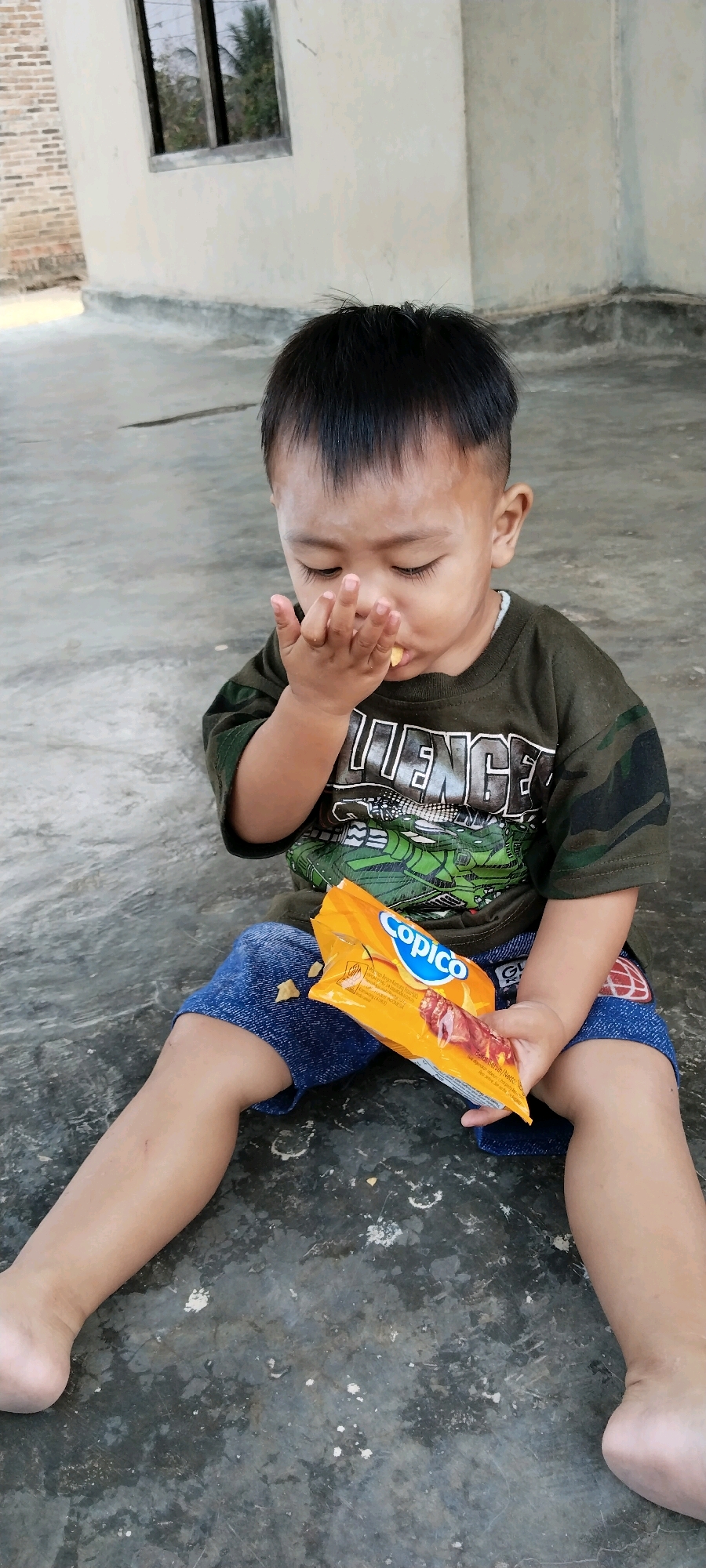 A young child with short black hair and dark eyes sits on a concrete floor, eating a snack. The child is wearing a green camouflage t-shirt and blue denim shorts with patches. In their right hand, the child holds an open orange package of Copico snacks, from which they are taking a bite. Several individual snacks are scattered on the floor around the child and on their shorts. The background consists of a beige wall with a window, and a brick wall to the left. The scene appears to be outdoors, possibly in the city of Tanjung Ratu Ilir, Indonesia, given the architectural style and vegetation visible through the window.