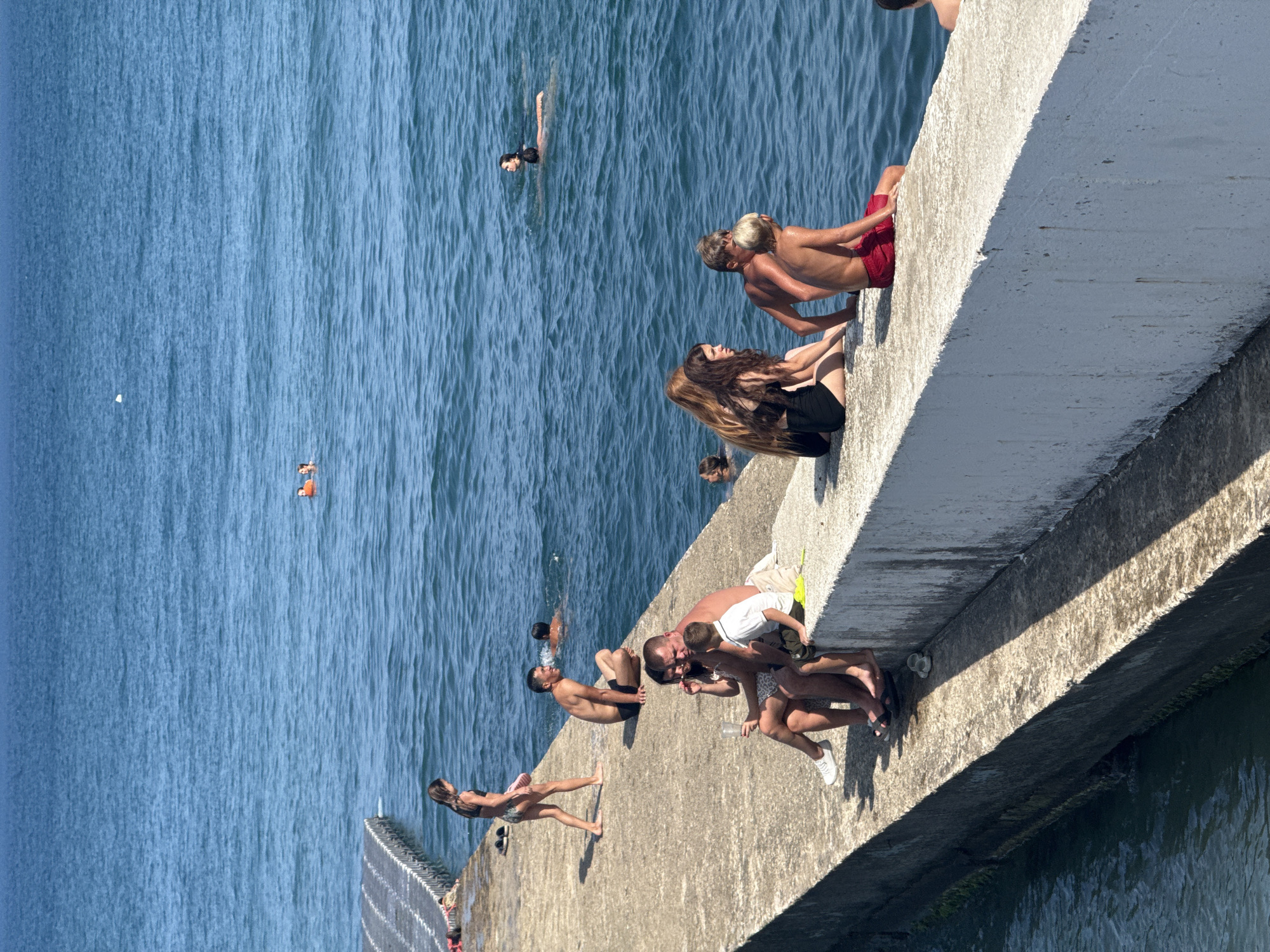 This media file depicts a group of people enjoying a sunny day by the sea on a concrete structure. The scene is set in Partenit, Ukraine, with the bright blue water indicating clear, pleasant weather. The time of day appears to be midday, suggested by the strong shadows cast by the sun.
Several individuals are engaged in various activities. In the water, some people are swimming and wading. On the concrete structure, which resembles a pier or a seawall, a number of people are sitting, conversing, and relaxing. One group is positioned closer to the camera, with a man in a white t-shirt interacting with a child, and another person sitting nearby. Further along the structure, a man in red shorts is leaning back, while a woman in a black swimsuit sits beside him. Another woman with long brown hair is also seated on the edge. In the distance, several other individuals can be seen in the water. The overall mood seems to be relaxed and leisurely, consistent with a summer day at the beach. There are no visible text elements in the image.