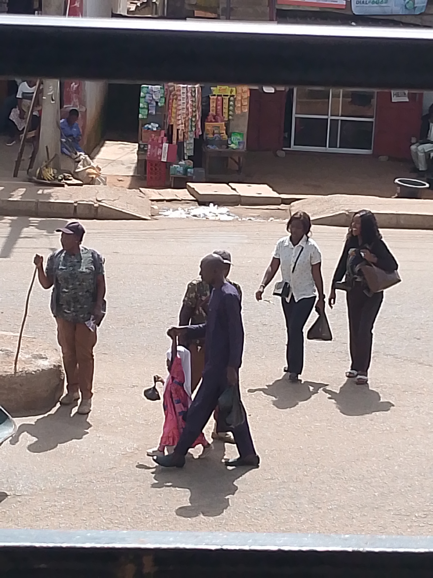 In Bukuru, Nigeria, a street scene unfolds with several individuals walking. In the foreground, a woman in a camouflage vest and brown pants walks with a stick, appearing to lead a group. She wears a baseball cap. Behind her, a man in a dark suit is seen from the waist down. To the right, two women walk together. The woman on the left wears a white patterned shirt and dark pants, carrying a black purse. The woman on the right is dressed in a black outfit and carries a brown bag. A young child in a pink dress is partially visible between the first woman and the man in the suit, holding onto something black. The background shows a row of shops and market stalls, suggesting a commercial area. The ground is paved with asphalt, and there are some raised concrete structures. The lighting indicates it is daytime.