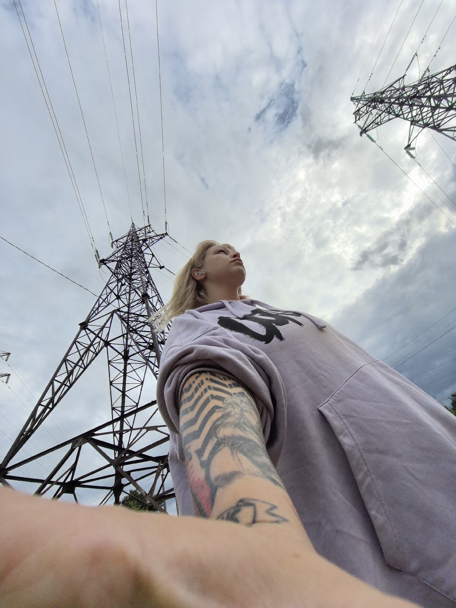 A low-angle outdoor shot captures a young person, likely female, with blonde hair, looking upwards towards a cloudy sky. She is positioned slightly to the right of the frame, wearing a light purple hooded sweatshirt with a stylized black graphic design on the chest. Her left arm, extended towards the camera, reveals a heavily tattooed sleeve featuring prominent zebra-like stripes on the upper arm and a smaller, abstract tattoo on her forearm.
The background is dominated by two towering metal electricity pylons, one central and one partially visible on the right, with numerous high-tension power lines stretching across the upper portion of the frame. The pylons are dark and intricate, showcasing their lattice-work construction and insulators. The sky is largely overcast with grey and white clouds, indicating a cloudy daytime setting. The composition emphasizes the scale of the industrial structures against the sky. The person appears to be posing for a photograph, looking contemplatively into the distance. The scene is set in an open area, typical of a power line corridor, near Noviy, Russia.