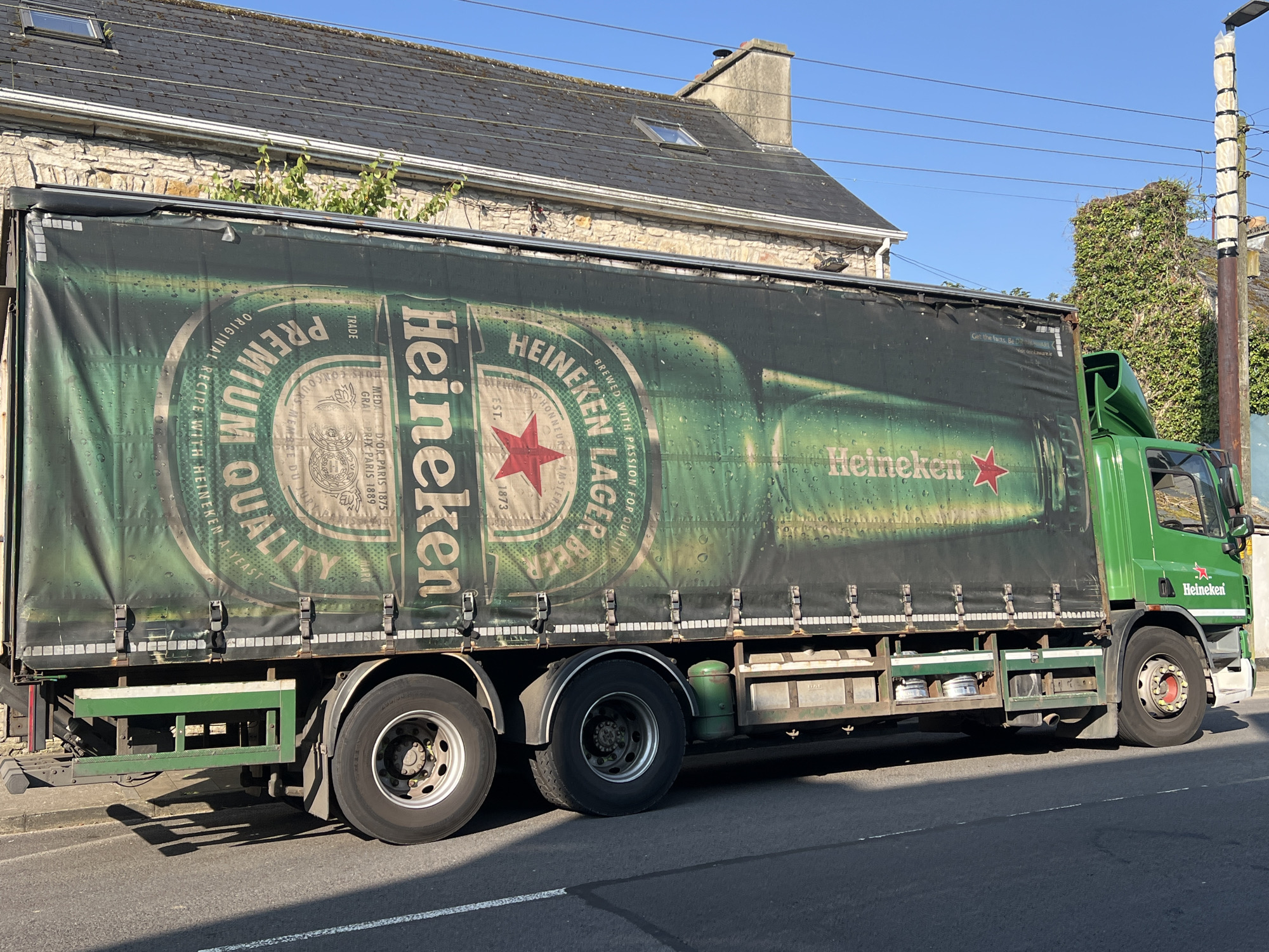 A green Heineken truck is parked on a paved road in front of a stone building with a grey slate roof. The truck's trailer is covered by a tarp featuring a large graphic of a Heineken bottle and branding. The truck is a right-hand drive vehicle, indicating its operation in Ireland. The background shows a clear blue sky and some greenery. The scene is well-lit by natural daylight, casting shadows from the truck onto the road.