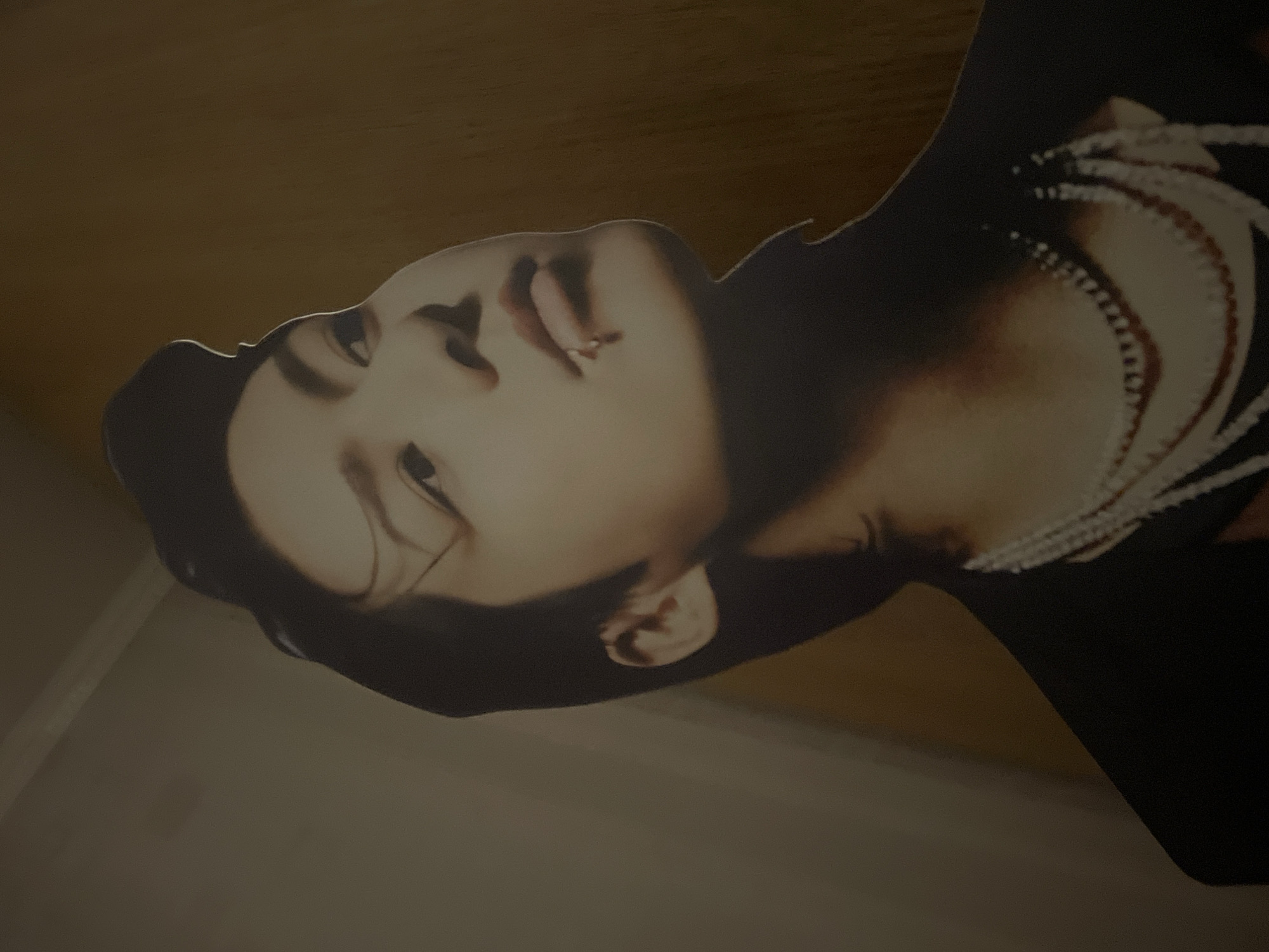 The image displays a tilted, close-up view of a cardboard cut-out featuring a person's head and upper torso. The individual depicted appears to be East Asian, with dark hair and a fair complexion. Their head is slightly tilted back, and they are looking upwards and to the viewer's right. Their expression is distinctive, with their tongue slightly extended and a silver lip piercing visible on their lower lip. They are wearing a dark-colored garment, possibly black, which features an elaborate white, possibly beaded or embroidered, design across the right shoulder and collarbone area.
The cut-out is set against a plain, warm-toned background, likely a wall, which has a slightly textured appearance. The lighting is somewhat dim, casting subtle shadows and highlighting the contours of the person's face. No specific activity is occurring; it is a static representation of an individual. There is no visible text in the image. The setting is contextually located in Saint Petersburg, Russia.
