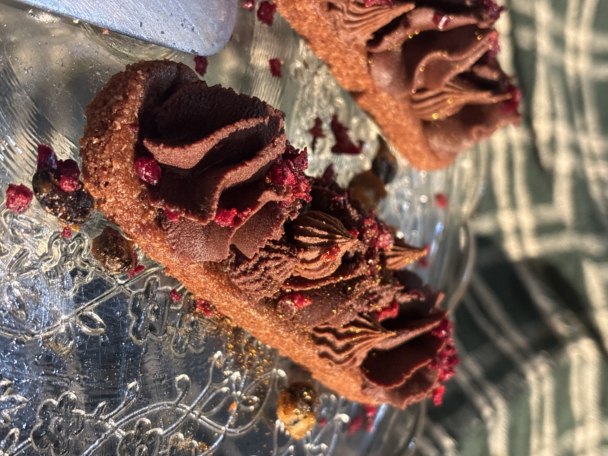 The image shows two small, oblong chocolate cakes with piped chocolate frosting and red berry crumble on top, sitting on a decorative clear glass plate. The cakes appear to be garnished with gold dust. The plate is set on a surface covered with a green and white checkered cloth. The lighting suggests the photo was taken outdoors or near a window with dappled sunlight. There are no people visible in the image. The location is not specified in the image itself and therefore cannot be determined as Riga, Latvia.