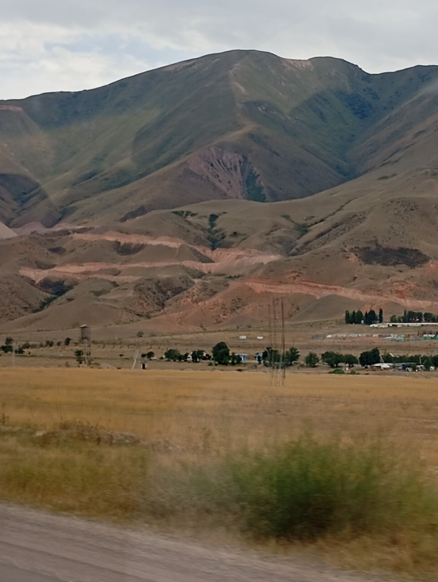 The image shows a vast, arid landscape dominated by rolling hills and mountains. In the foreground, dry, golden grass covers the ground, with a dirt road on the left edge. In the middle ground, a cluster of low-lying buildings and trees are visible, suggesting a small settlement. A water tower stands on the left side of the settlement. The background is characterized by large, barren hills and mountains, with some showing signs of erosion or mining activity, indicated by reddish-brown scars on their slopes. The sky is overcast with grey clouds. This scene is consistent with the landscape found in and around the city of Cholok, Kyrgyzstan, known for its mountainous terrain and dry climate.