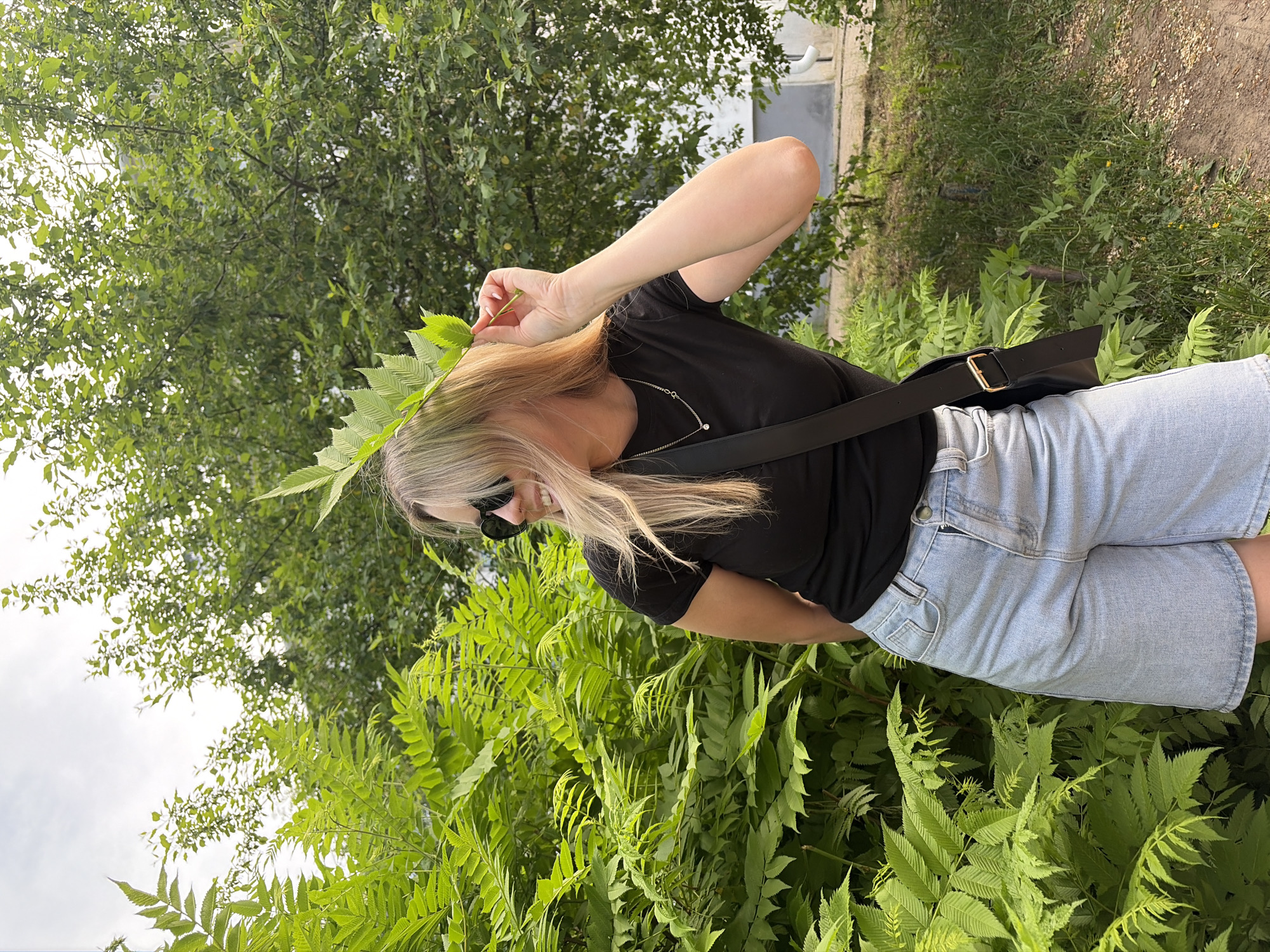 A woman is standing outdoors in a park-like setting filled with lush green foliage. The location is likely in Chaikovsky, Russia. She has long blonde hair, is wearing sunglasses, a black t-shirt, and light-colored denim shorts. A black shoulder bag is slung across her body. She is holding a leafy branch up near her head. The environment includes tall trees and dense green ferns.