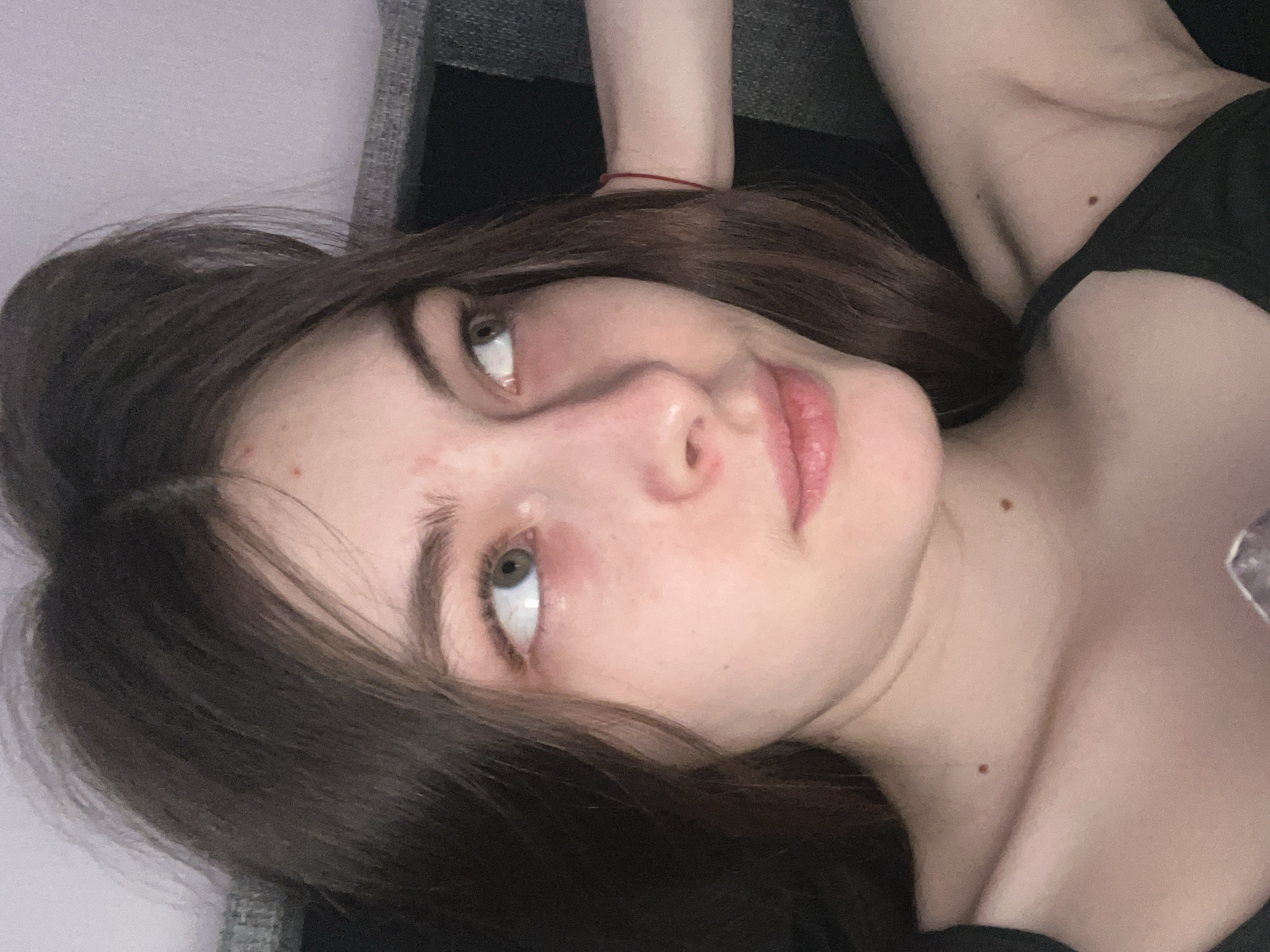 A young woman with brown hair and fair skin lies on a gray surface, likely a sofa or bed. Her head is tilted to the side, and she looks upward. She has light green eyes, and there are a few minor skin blemishes on her face. She is wearing a dark-colored top. A red string bracelet is visible on her wrist. Two moles are located on her neck and chest. The background includes the gray surface she is lying on and a dark, undefined area, potentially another part of the furniture. The location in the city of Saint Petersburg, Russia is unknown.