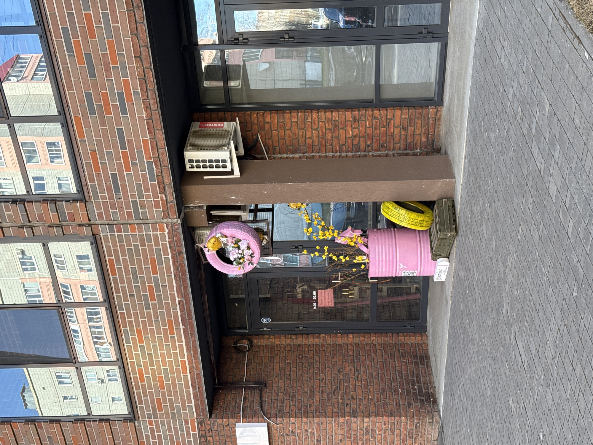 The image shows the exterior of a building, likely a commercial space in Izhevsk, Russia. The building has a brick facade with several windows reflecting nearby structures. An air conditioning unit is mounted near the top windows. The ground floor features a glass door and window arrangement with pink and yellow painted tires and a barrel used as decorative planters. A sign is visible near the planter. The sidewalk is paved with gray bricks.