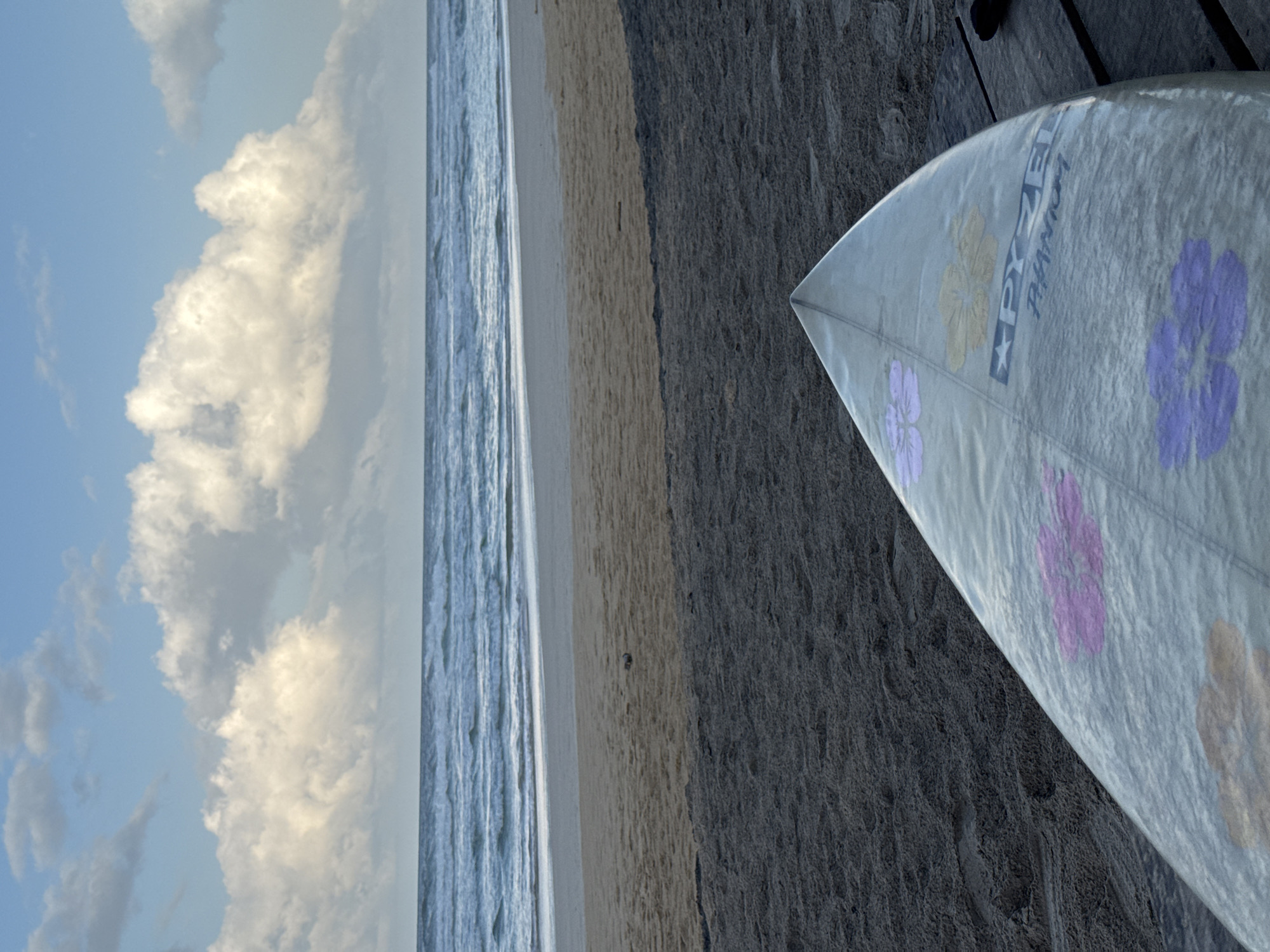 The image shows a surfboard lying on a sandy beach in Kathaluwa, Sri Lanka. The surfboard is light-colored with colorful floral designs and brandings. The beach transitions from dark sand in the foreground to lighter sand further back, with the ocean and waves visible in the distance under a cloudy sky. The scene is tranquil and captures a typical coastal landscape.