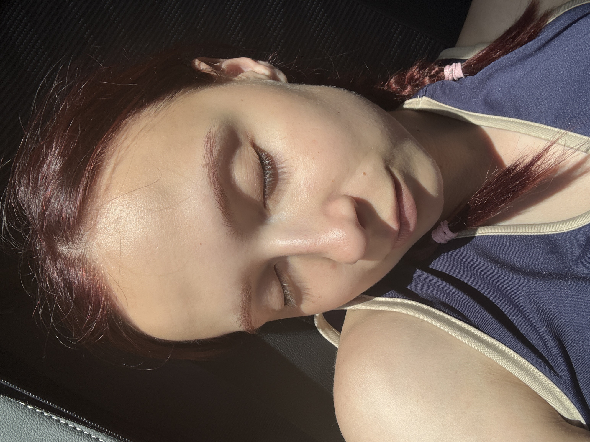 A close-up, high-angle shot depicts a fair-skinned person with burgundy-toned hair styled into braids secured with pink hair ties. The person is reclined, eyes closed, resting on a dark surface, possibly a car seat. They are wearing a navy blue top with a light beige trim. The lighting suggests an indoor setting with sunlight present. There is no verifiable indication the location is Можга, Russia.