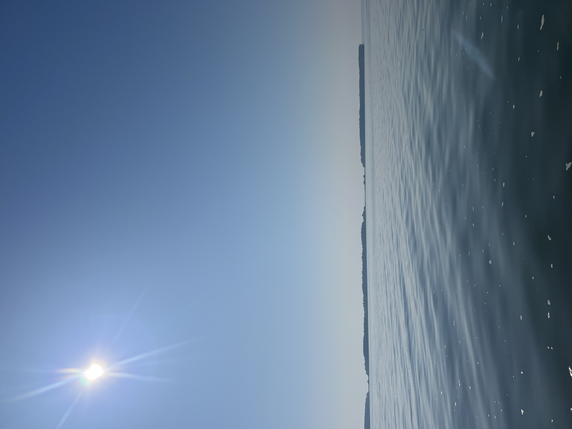 The image shows a serene body of water under a clear blue sky. The sun, positioned in the upper left corner, is bright and casts visible rays. In the distance, several elongated islands covered in dark green foliage dot the horizon. The water's surface is calm with gentle ripples and a scattering of small white flecks, possibly foam or debris. There are no people or man-made structures visible in the frame, except for what could be a very faint line extending from the islands on the left. The scene is likely a natural landscape with islands in a lake or sea.