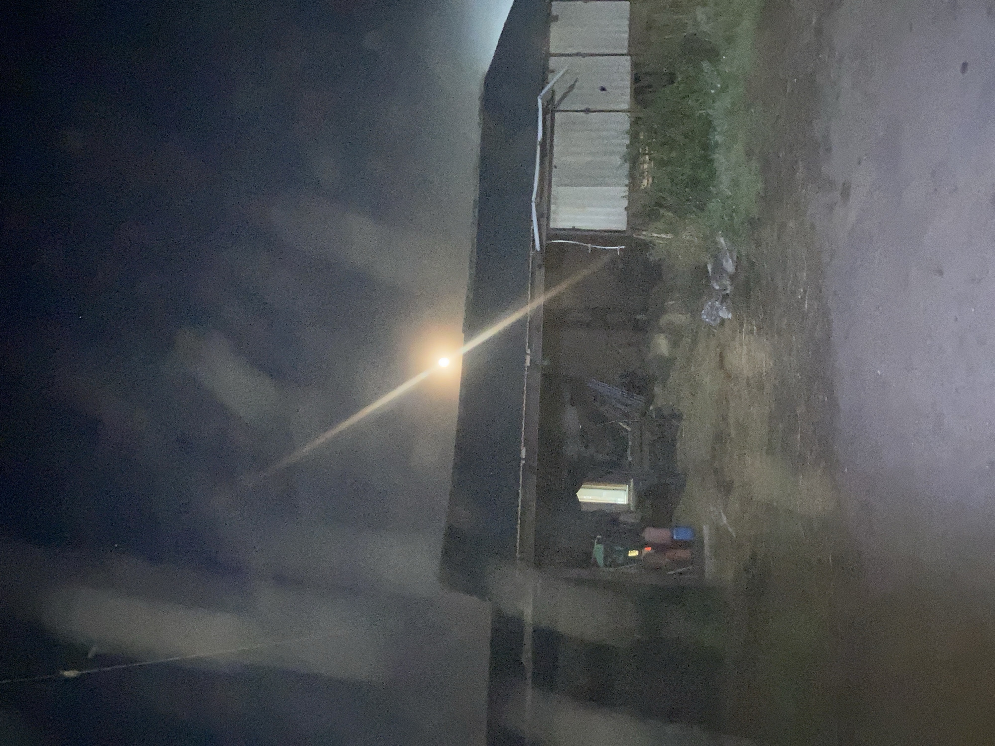 The image depicts a nighttime scene in the city of Besedy, Russia, featuring a rustic barn with a dark roof. A bright, indistinct light source, possibly the moon, is visible above the barn, casting a strong beam of light that cuts across the frame. The barn's exterior appears weathered, with some sections covered in corrugated metal siding. In front of the barn, there is a dirt path and overgrown vegetation. To the left of the barn, two figures, a male and a female, are visible. The male is wearing a light-colored shirt, and the female is wearing a darker garment. They are standing near a blue bucket and some other unspecified objects. The overall atmosphere is dimly lit and somewhat hazy, possibly due to atmospheric conditions or light diffusion.