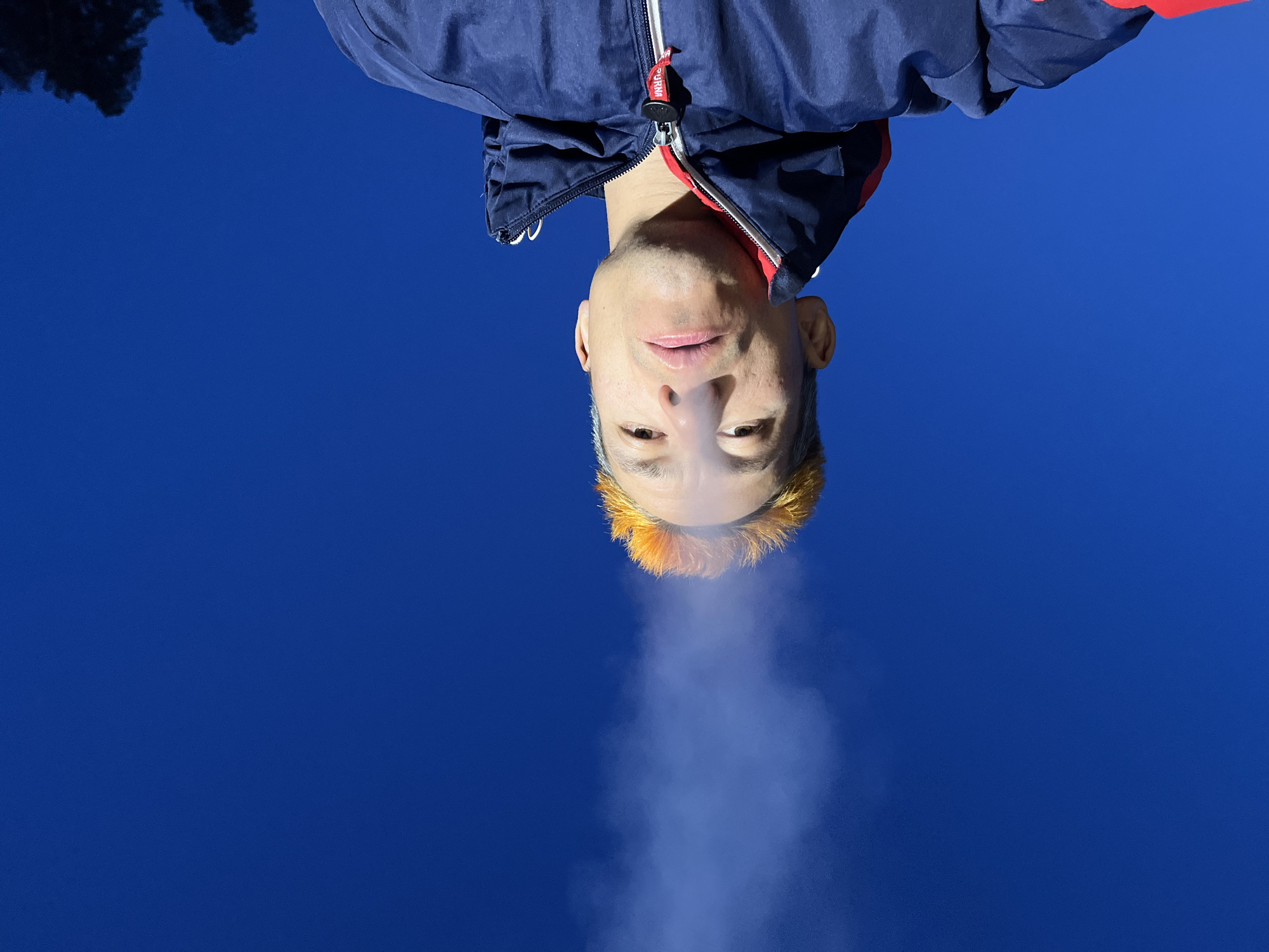 This media file presents a striking low-angle portrait of a man, appearing inverted against a vibrant, cloudless deep blue sky. The man is captured from the chest up, looking directly down (or up, depending on orientation) towards the camera. He wears a blue jacket with red accents and a silver zipper, on which a red and white tag featuring the brand name "PUMA" is visible. His short hair, closest to the bottom of the frame, is dyed a distinct orange or yellow, fading into a lighter shade further up his head. He has a light mustache and stubble, and his expression is neutral.
A column of white vapor or smoke is exhaled from his mouth, rising into the sky, suggesting either vaping or visible breath in cold air. In the upper left corner of the frame (which would be the lower left if rotated), the dark silhouettes of distant tree branches are barely visible against the blue, indicating an outdoor setting. The clear sky and visible breath imply a cold, sunny daytime. The unusual perspective creates a visually dynamic and slightly surreal composition, possibly set in Chaikovsky, Russia.