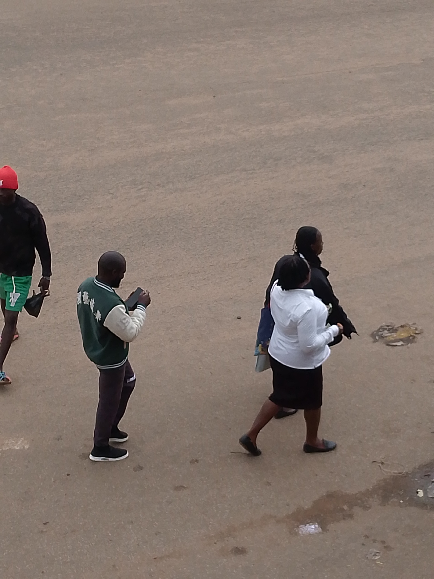 The image depicts a street scene in Bukuru, Nigeria. Four individuals are visible. On the left, a person wearing a red cap and a black jacket walks away from the viewer, carrying a black bag. To their right, a person wearing a green and white jacket is looking at and holding a smartphone. Further to the right, two people are walking together. One is wearing a white shirt and a black skirt, and the other is wearing a black outfit. They are walking towards the right of the frame. The ground is paved and appears to be a road or sidewalk, with some debris scattered on the surface. The overall lighting suggests an overcast day.