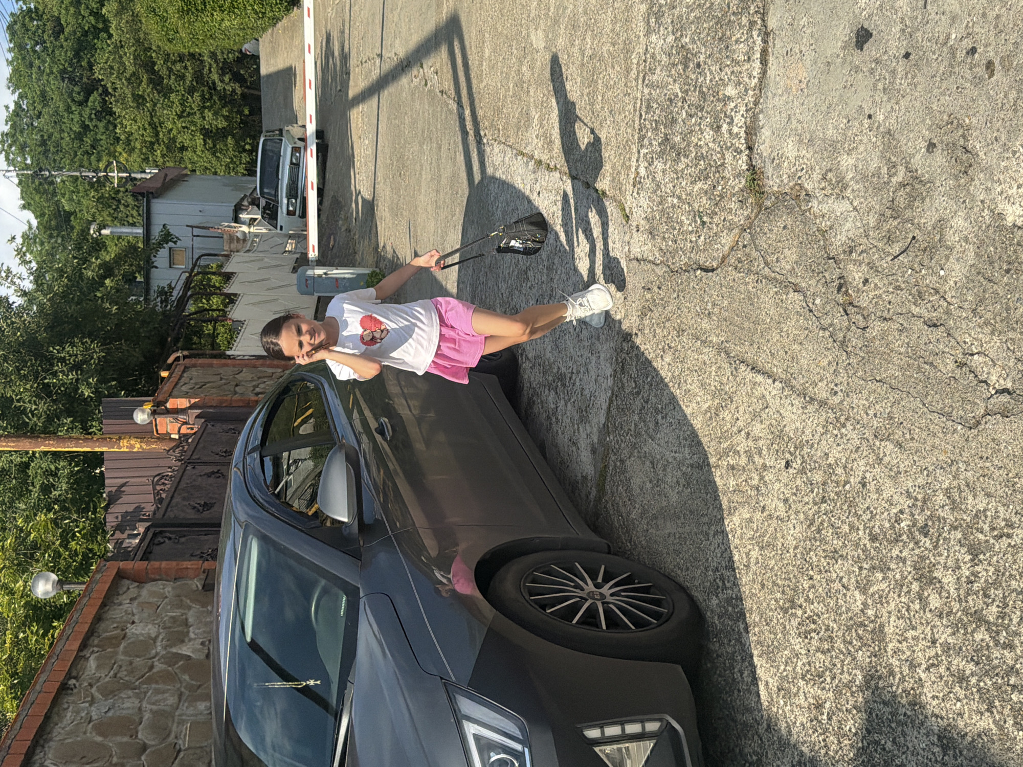 In Sochi, Russia, on a sunlit day, a young girl with dark hair, wearing a white t-shirt with a teddy bear graphic and a pink skirt, walks on a cracked asphalt path. She holds a small black handbag and is on her phone, seemingly mid-conversation. The path leads past a dark grey sedan parked on the left side of the frame, with a stone wall and ornate iron gate in the background. Beyond the gate, trees line the upward sloping road, where a white car is visible near a barrier. The scene is bathed in warm sunlight, casting long shadows across the ground.