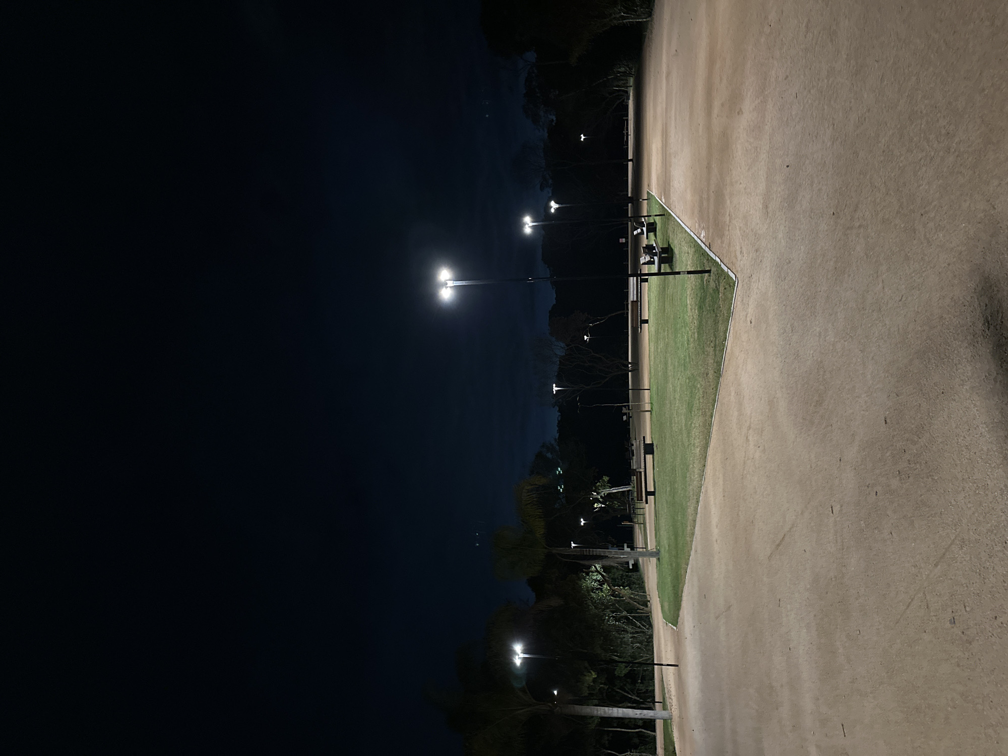 This is a nighttime photograph of an empty park area with paved walking paths and a grassy area. Several lampposts are illuminating the scene. In the background, there are trees and some distant lights. There are benches on the grassy area. The image does not depict any people or actions. The location is specified as Florianópolis, Brazil, and the scene appears to be a public park or recreational area within the city.